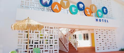 Un hotel llamado 'WATERSIDE' con fachada blanca, una escalera que lleva a la entrada y mesas exteriores con sillas y un paraguas de paja.