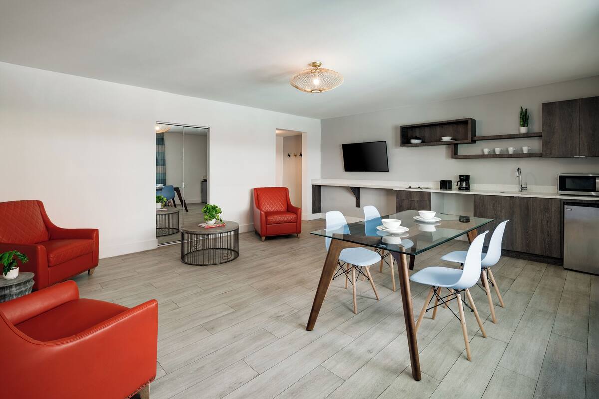 A modern living room with a glass dining table, red and white chairs, and a red armchair. A kitchen area with a TV, cabinets, and a microwave is visible in the background.