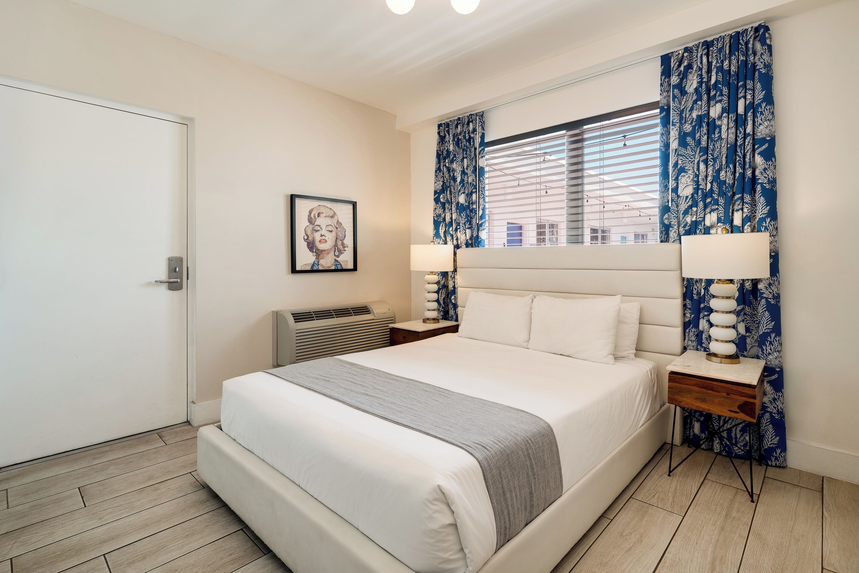 A modern hotel room with a large bed, a bedside table with a lamp, a window with blue curtains, and a framed picture on the wall.