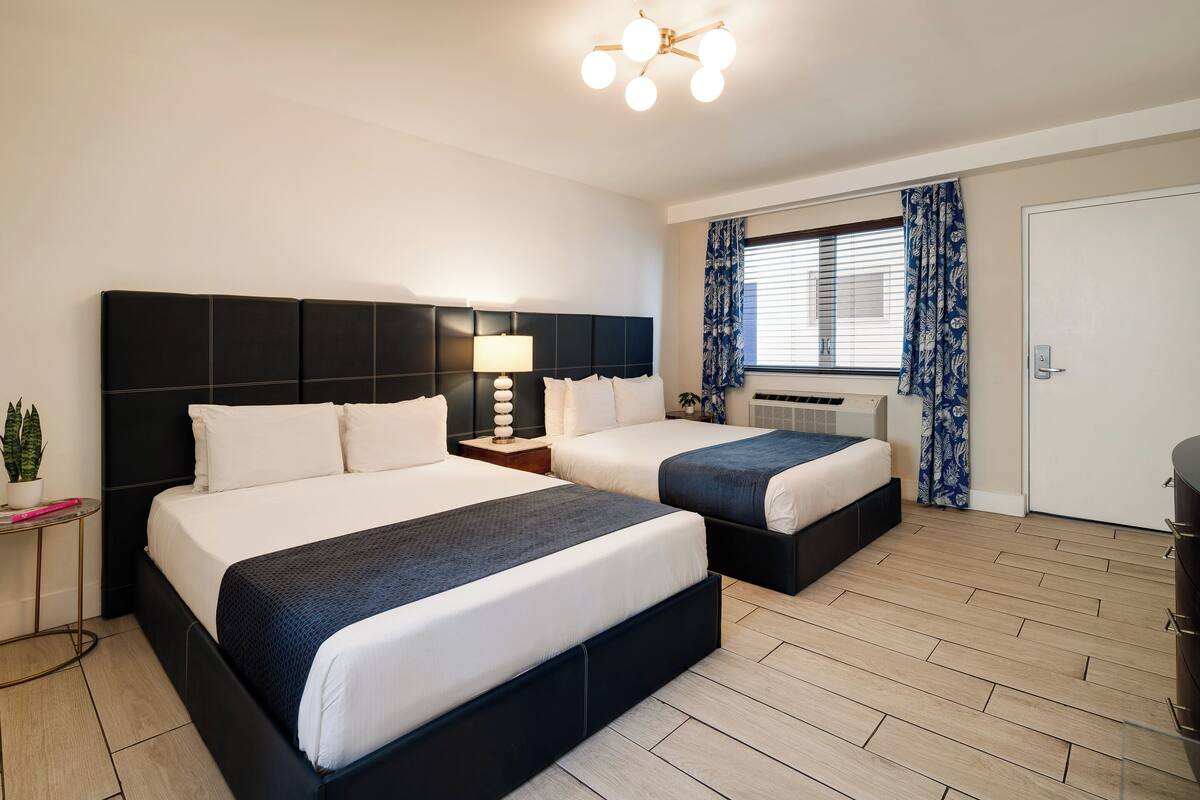 A double room with two beds, a bedside table with a lamp, a window with curtains, and a door.