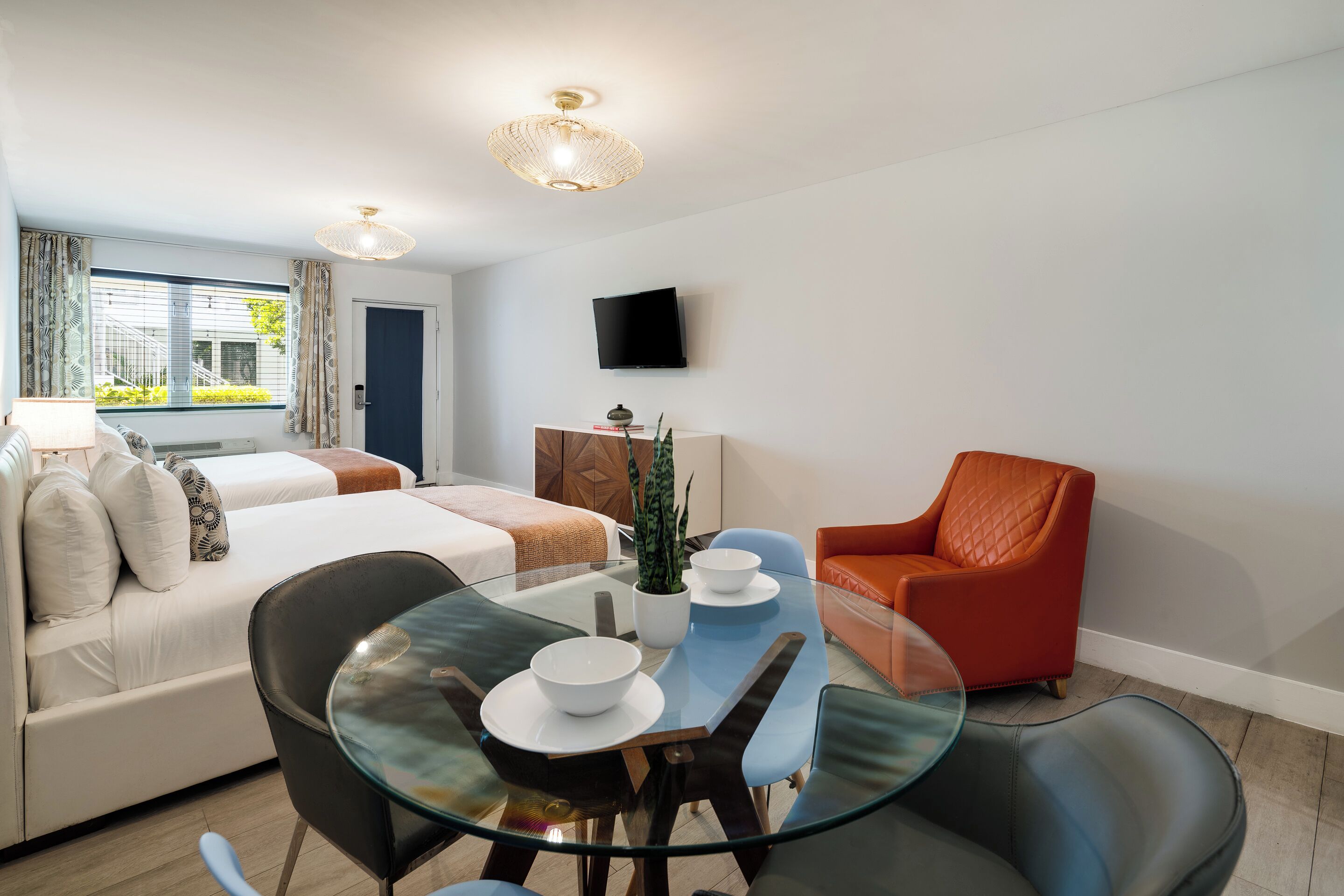 A modern hotel room with a glass dining table, a sofa, a red armchair, a TV mounted on the wall, and a window with curtains.