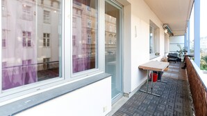 A balcony with a table and chairs, a red bucket, and a view of buildings through large windows.