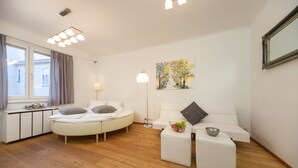 A modern, well-lit bedroom with a bed, a sofa, and a small table. There is a painting on the wall and a window with curtains.