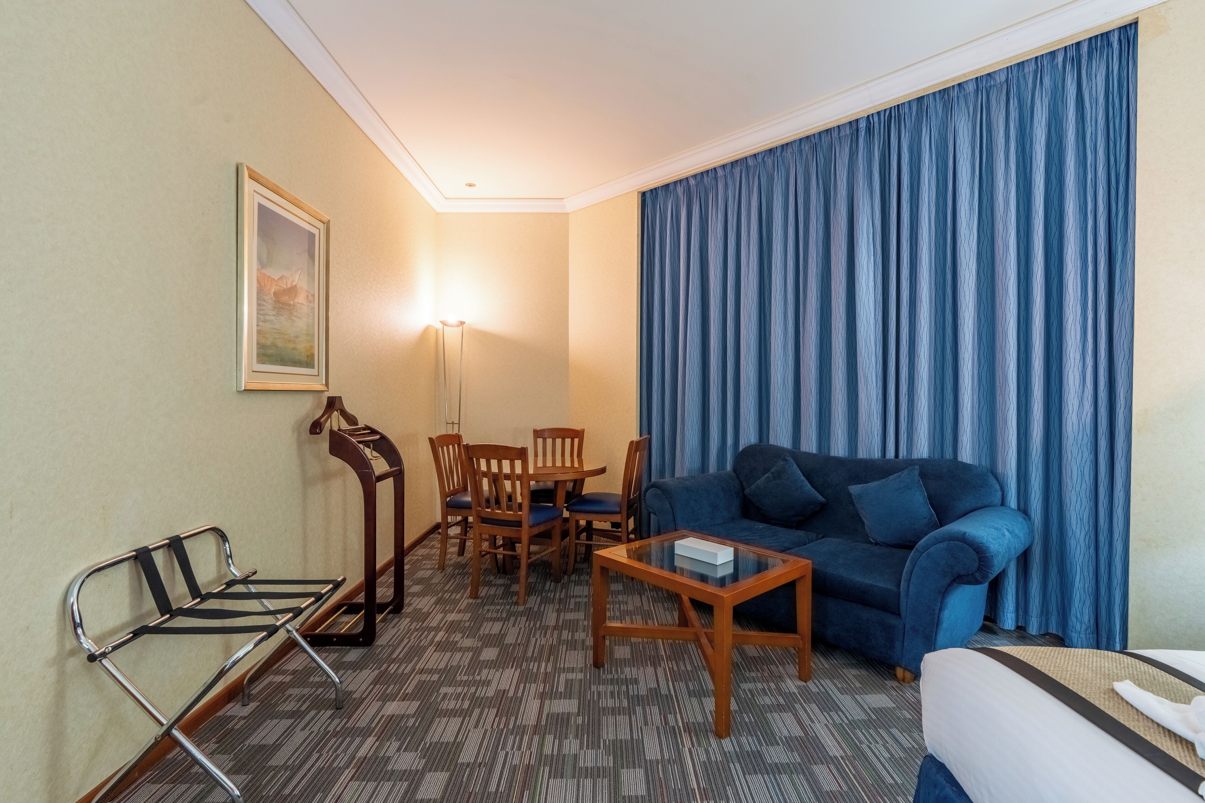 Et hotelværelse med en blå sofa, et træspisebord med stole, et lille træsofabord, en metalbænk og et stort vindue med blå gardiner.