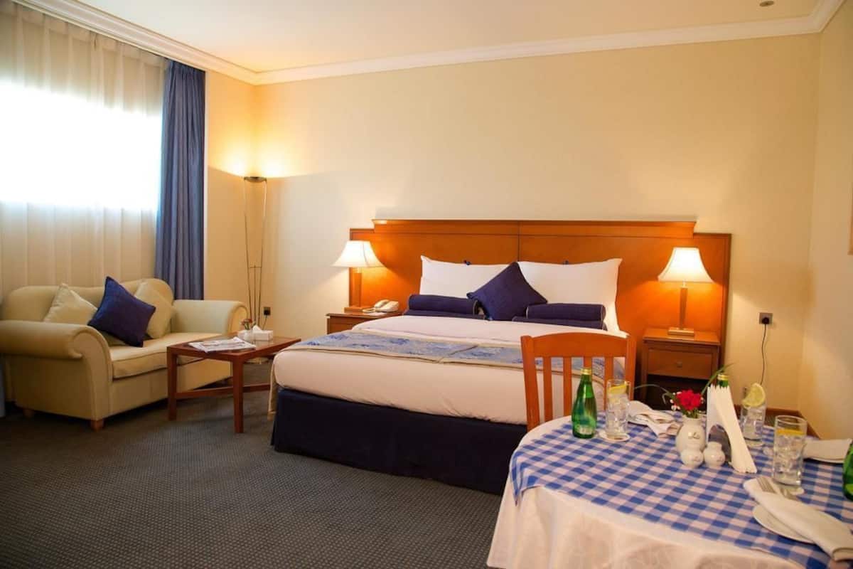 Une chambre d’hôtel avec un lit, des tables de chevet, une chaise, une petite table recouverte d’une nappe à carreaux et une fenêtre avec des rideaux.