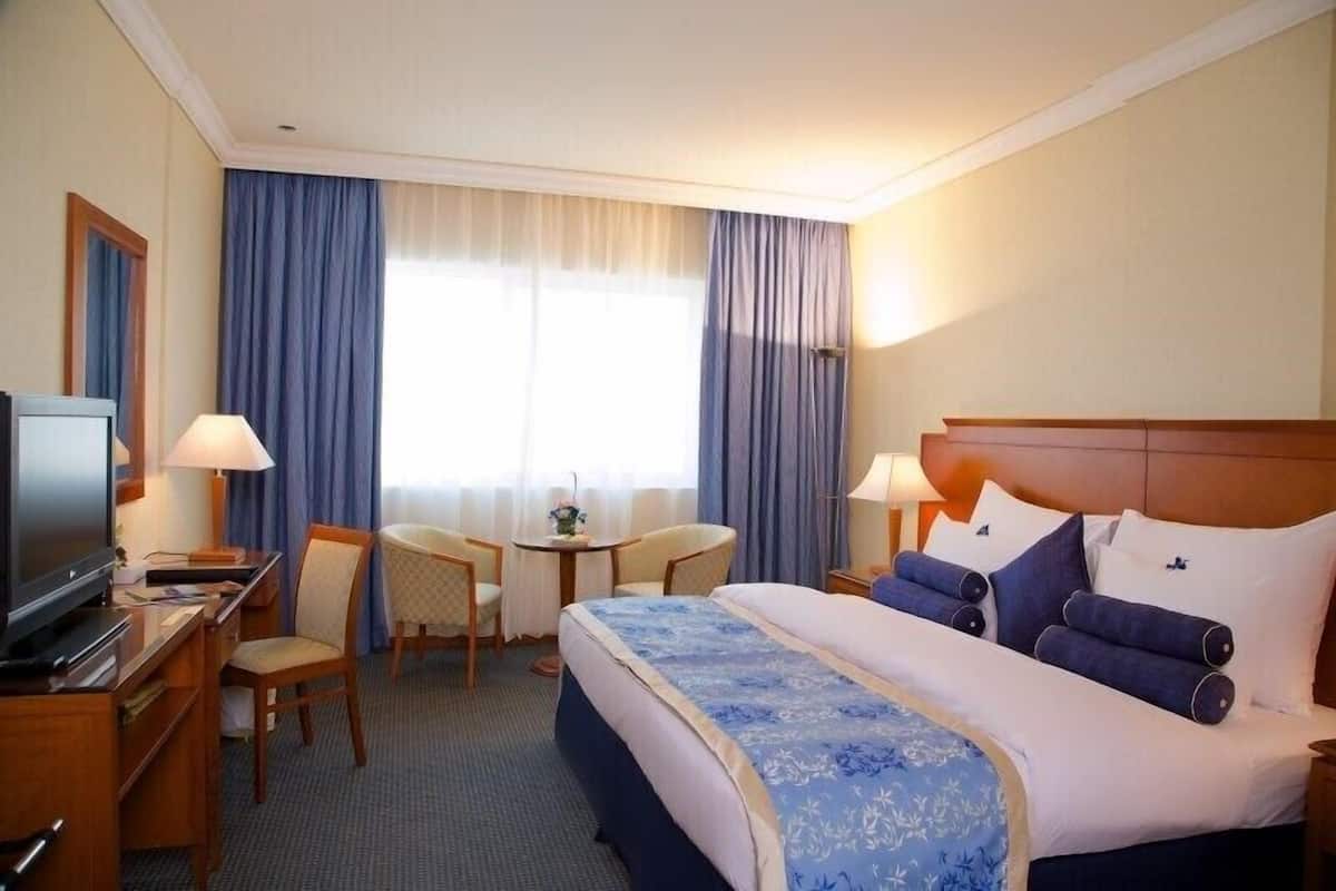 Une chambre d’hôtel avec un lit, un bureau avec une télévision, une chaise, une petite table avec une fleur et une fenêtre avec des rideaux.