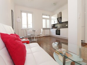 A modern living room with a white sofa, red pillow, and glass coffee table. There is a dining area with a white table and chairs, and a kitchen with white cabinets and a black stove. - Vienna Apartment Center - Zentrum I (Vienna)