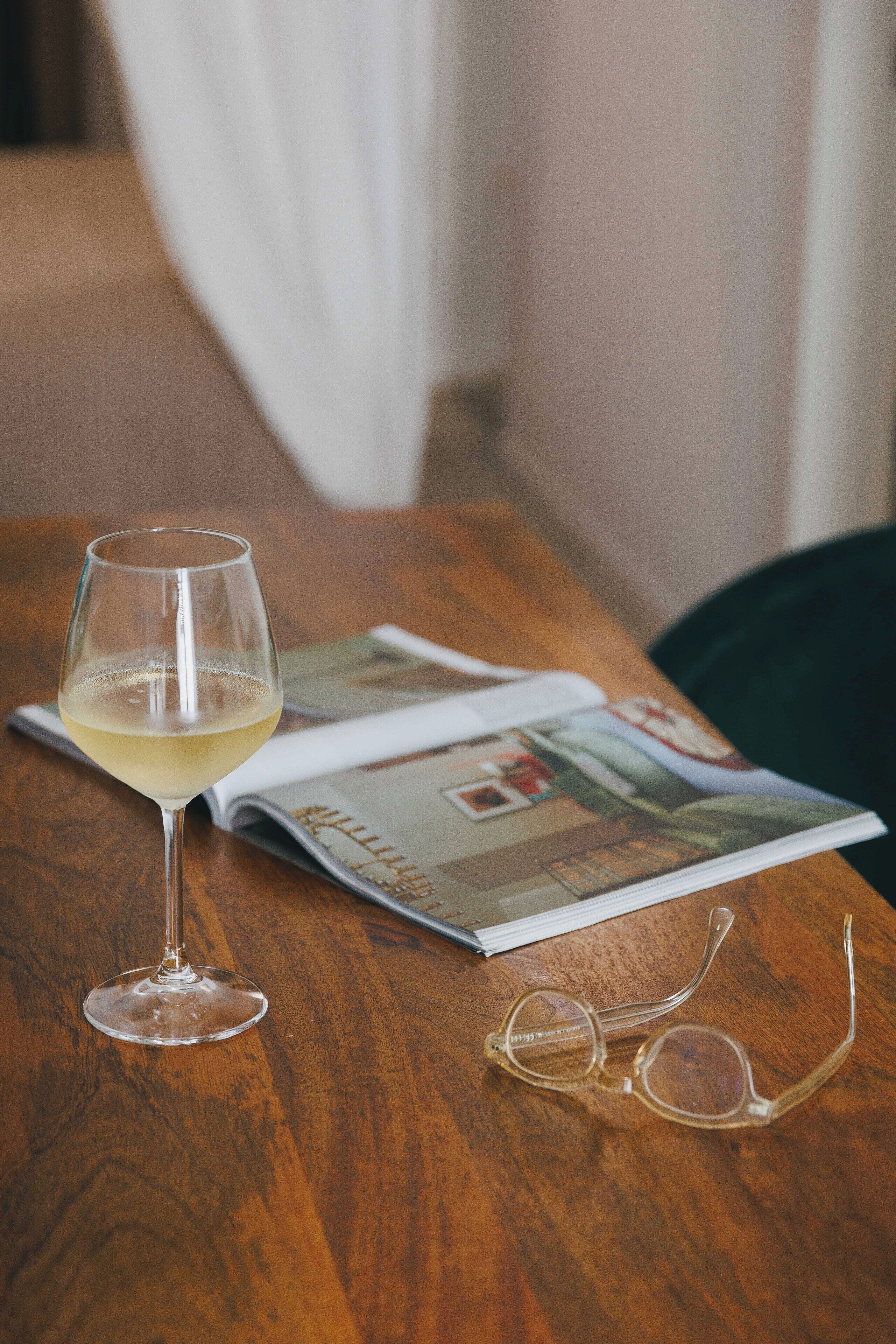 Una mesa de madera con una copa de vino, un par de gafas y una revista abierta.