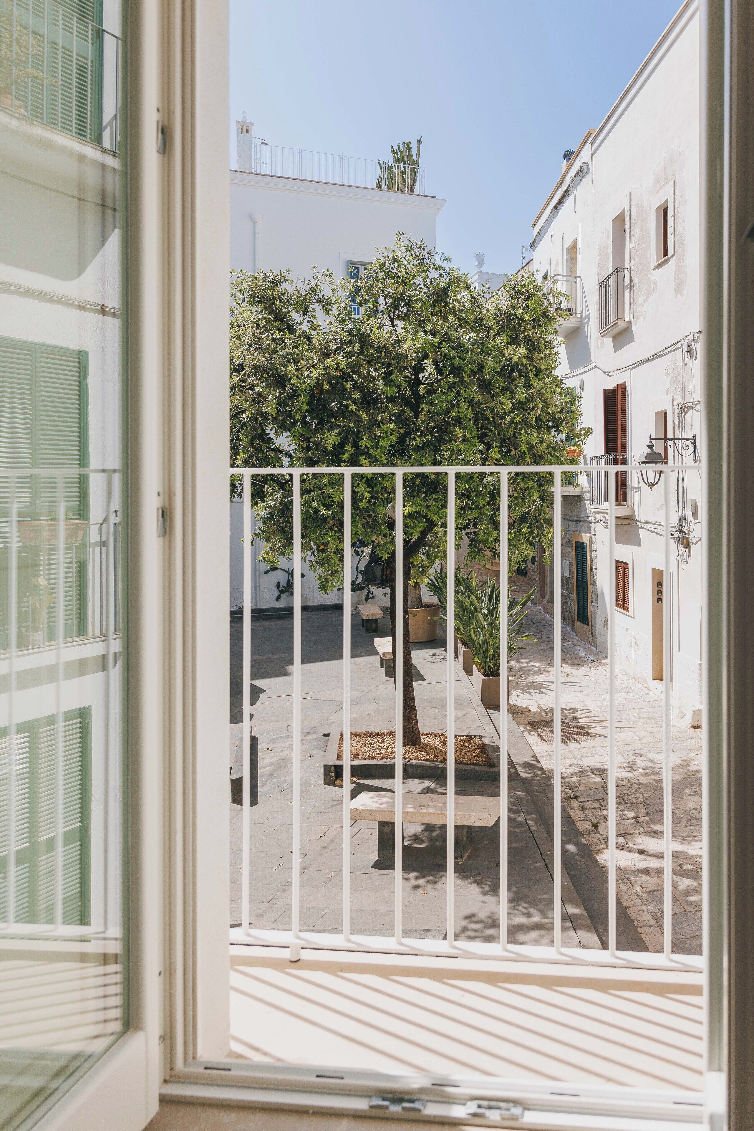 Vista desde un balcón con baranda de metal blanco, sobre una calle estrecha con edificios y un árbol.