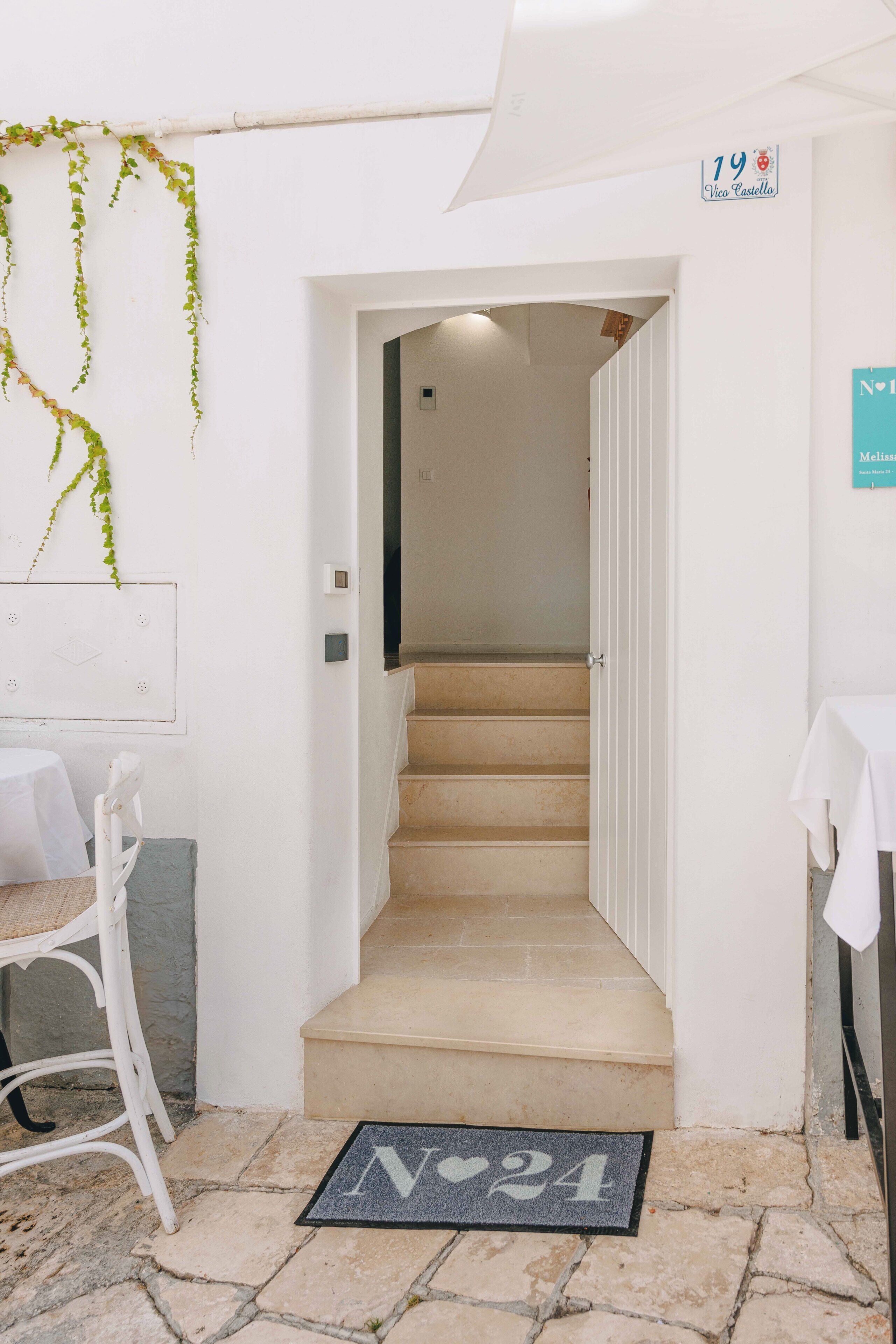 Eine Treppe führt zu einer oberen Etage, ein weißer Stuhl, eine Fußmatte mit der Aufschrift 'No 24' und ein Schild mit '19 Vico Castello'.