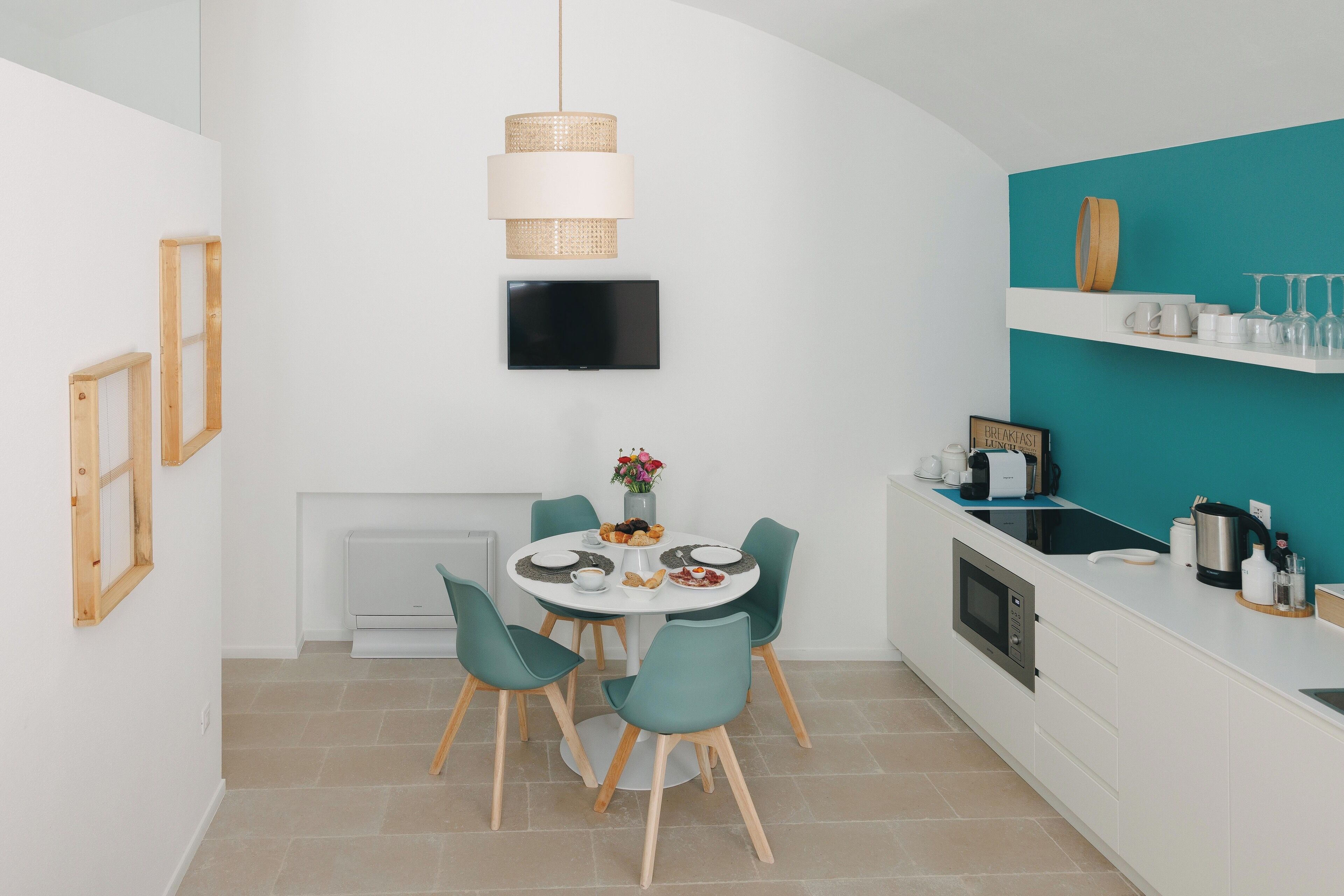 A modern kitchen with a dining area, white cabinetry, a teal accent wall, a flat-screen TV, and a hanging light fixture.
