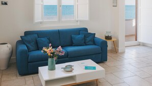 A modern living room with a blue sofa, a white coffee table, and a view of the sea through a large window. - Santa Maria 24 - Luxury Apartments (Monopoli)