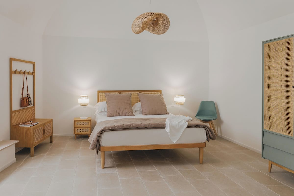 Une chambre avec un lit en bois, des tables de chevet, une suspension en osier, un fauteuil vert et une armoire en rotin.