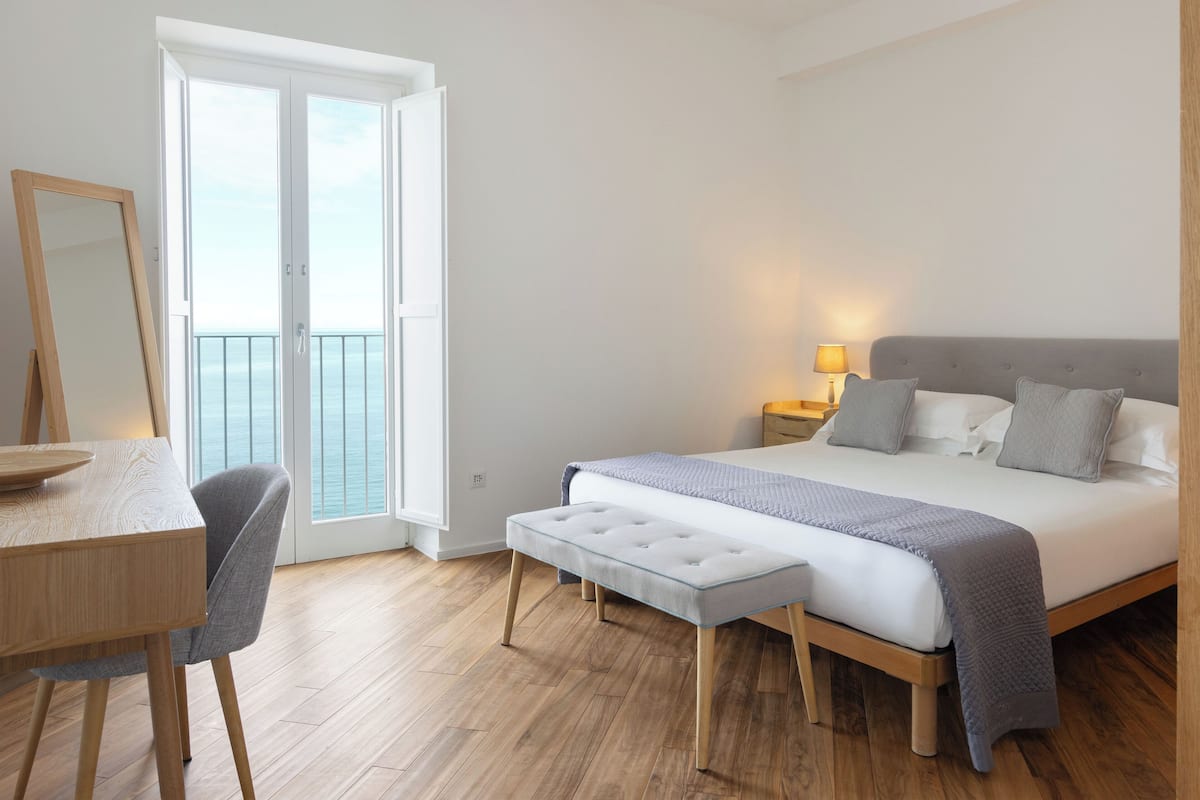 Une chambre à coucher avec un lit, une table de chevet, une lampe et une vue sur la mer, visible par une porte.