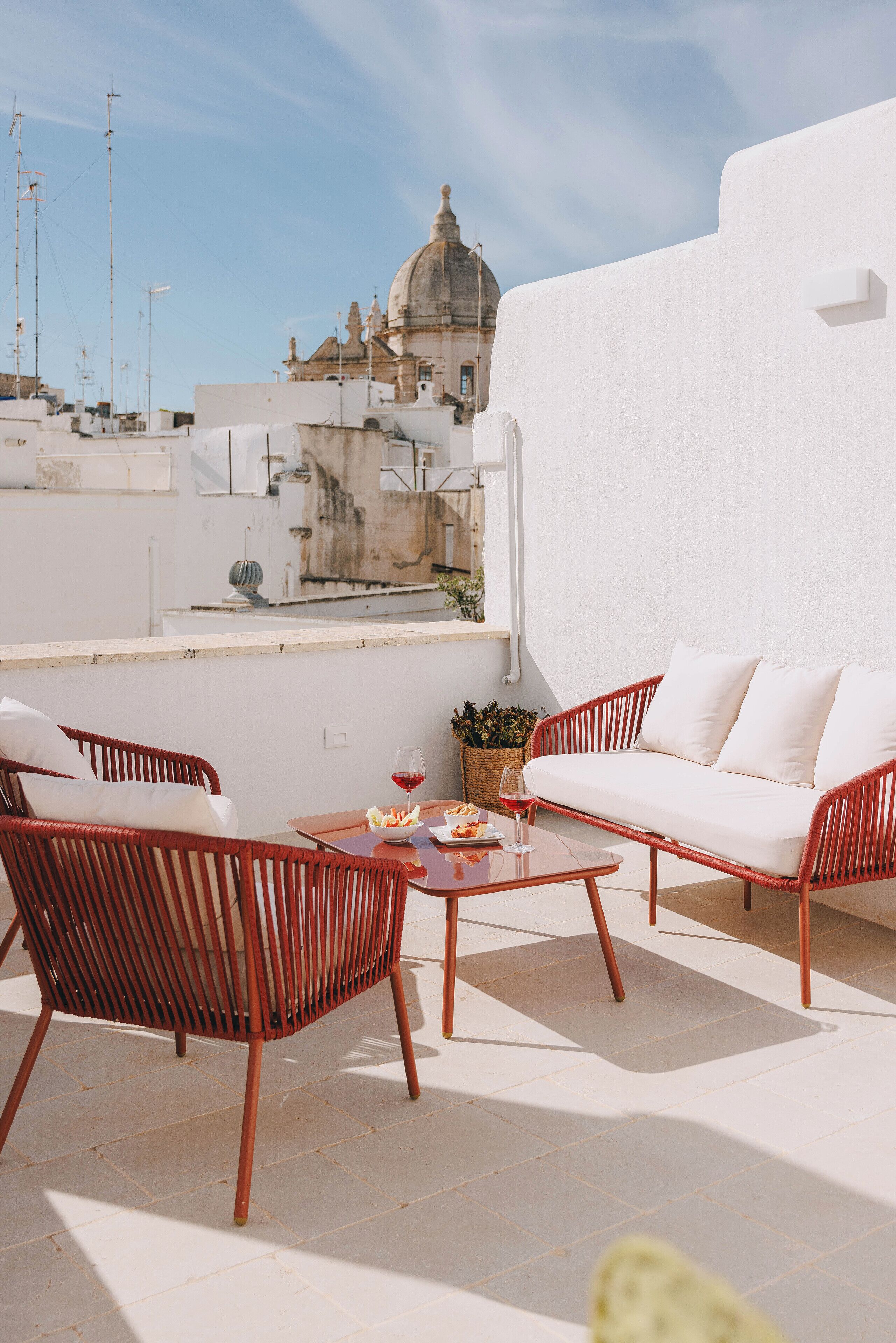 A rooftop terrace with a white sofa, red wicker chairs, and a table with a basket and glass of wine.