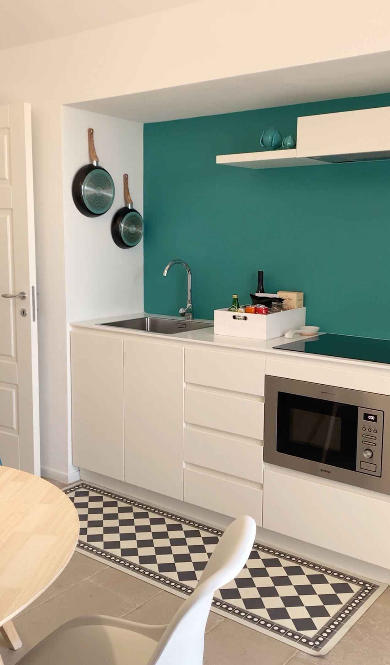 A compact kitchen with white cabinets, a teal backsplash, a sink, a microwave, and a checkered floor mat.