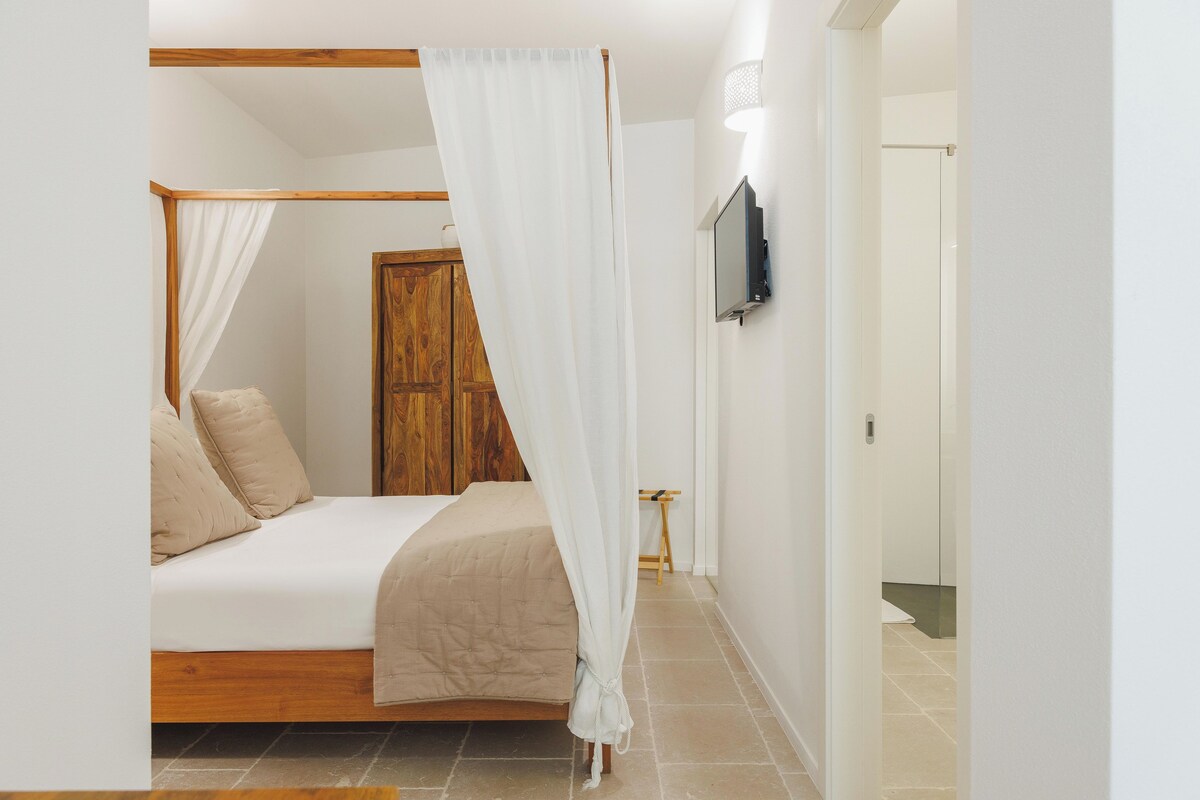 Un dormitorio con cama, cabecera de madera, dosel blanco y televisión en la pared.