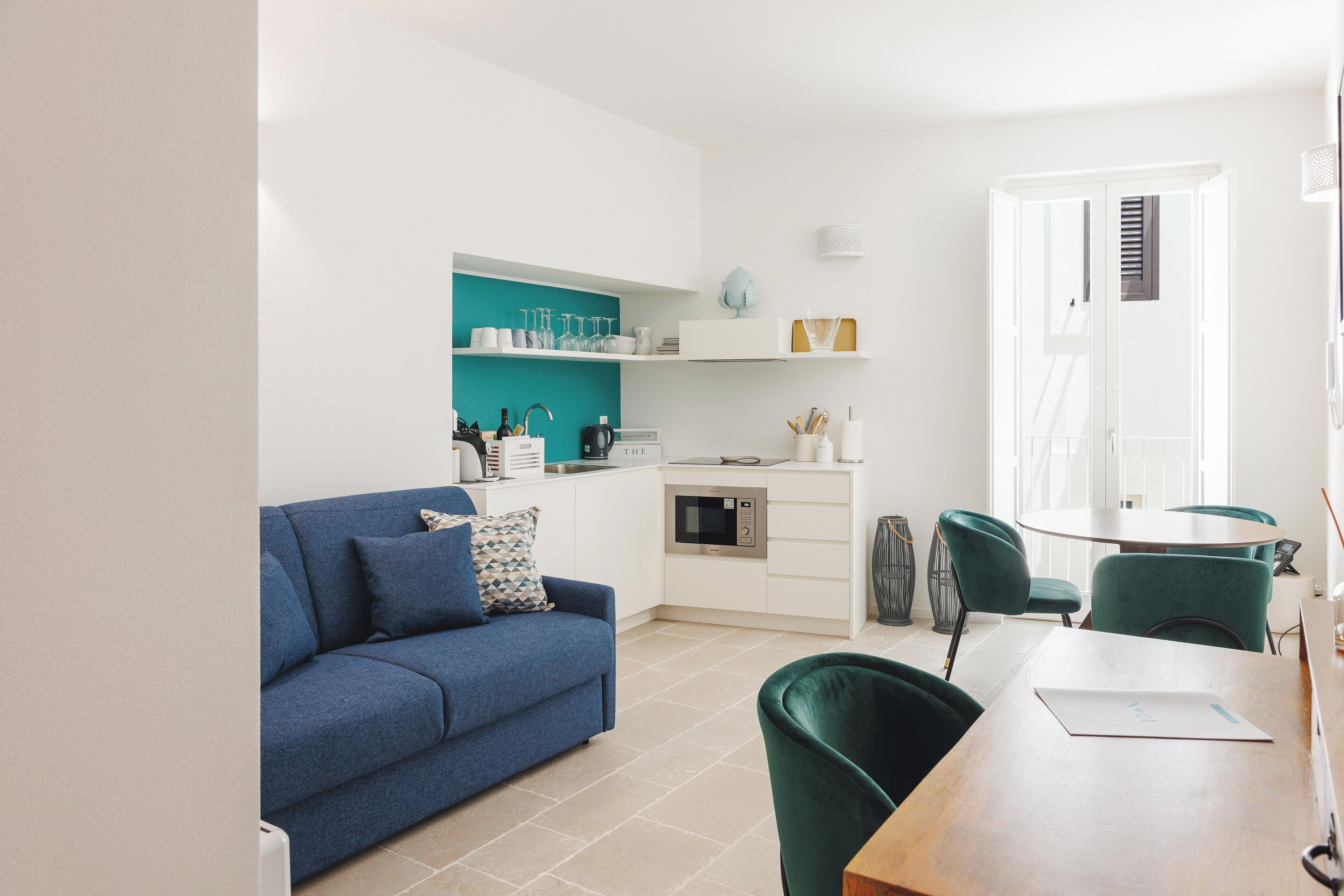 A modern living room with a blue sofa, a wooden dining table, and a kitchen area with white cabinets and teal shelves.