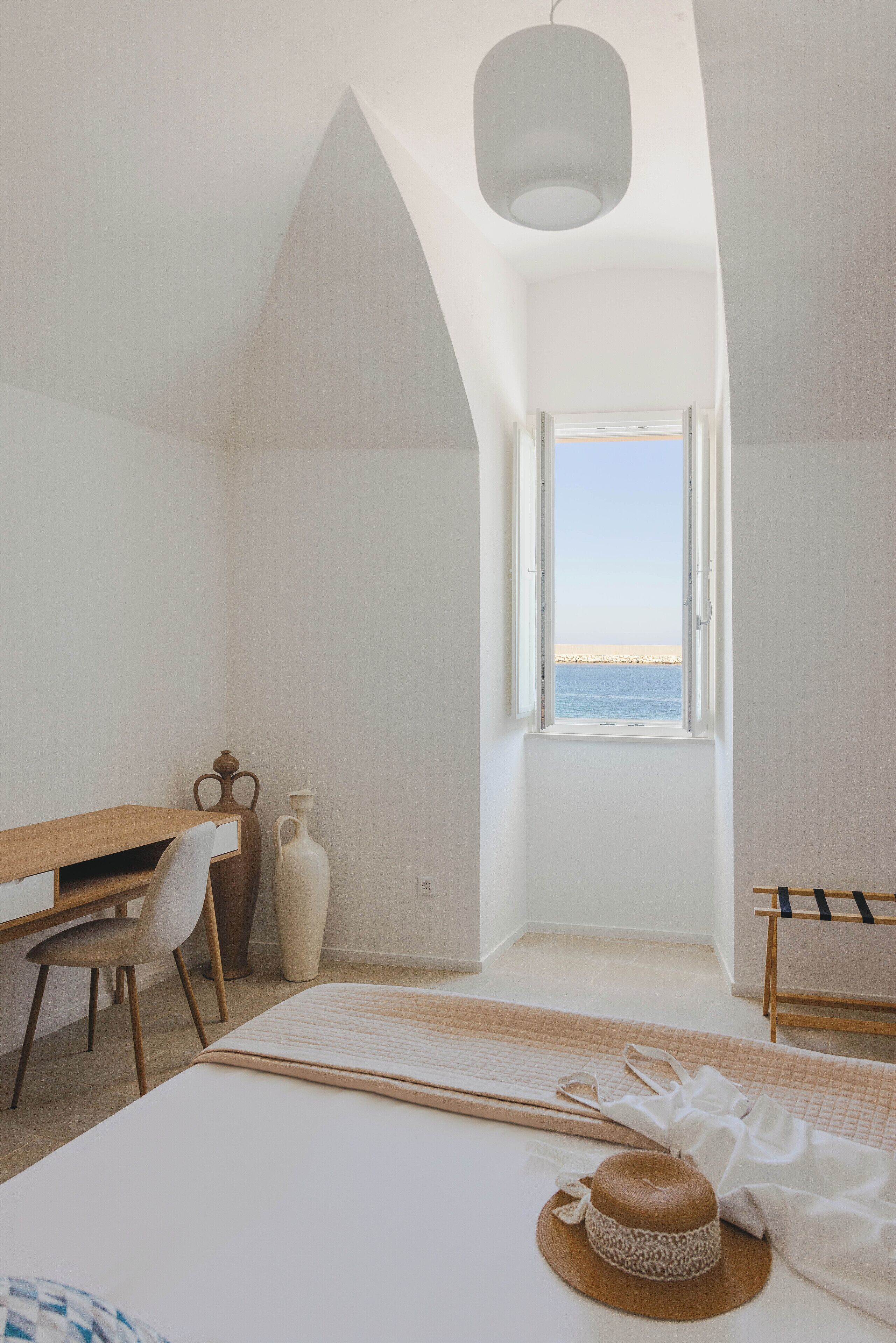 A bedroom with a bed, a desk, a chair, a window with a view of the sea, and a hanging light fixture.