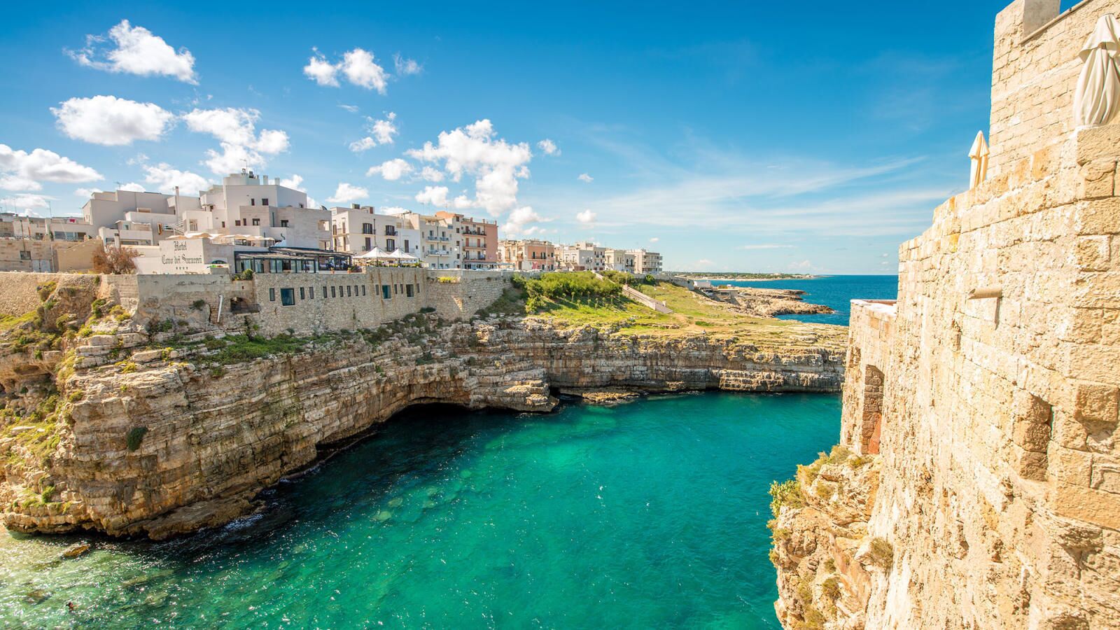 Une ville côtière avec des bâtiments en pierre, une mer d’un bleu limpide et une falaise rocheuse.