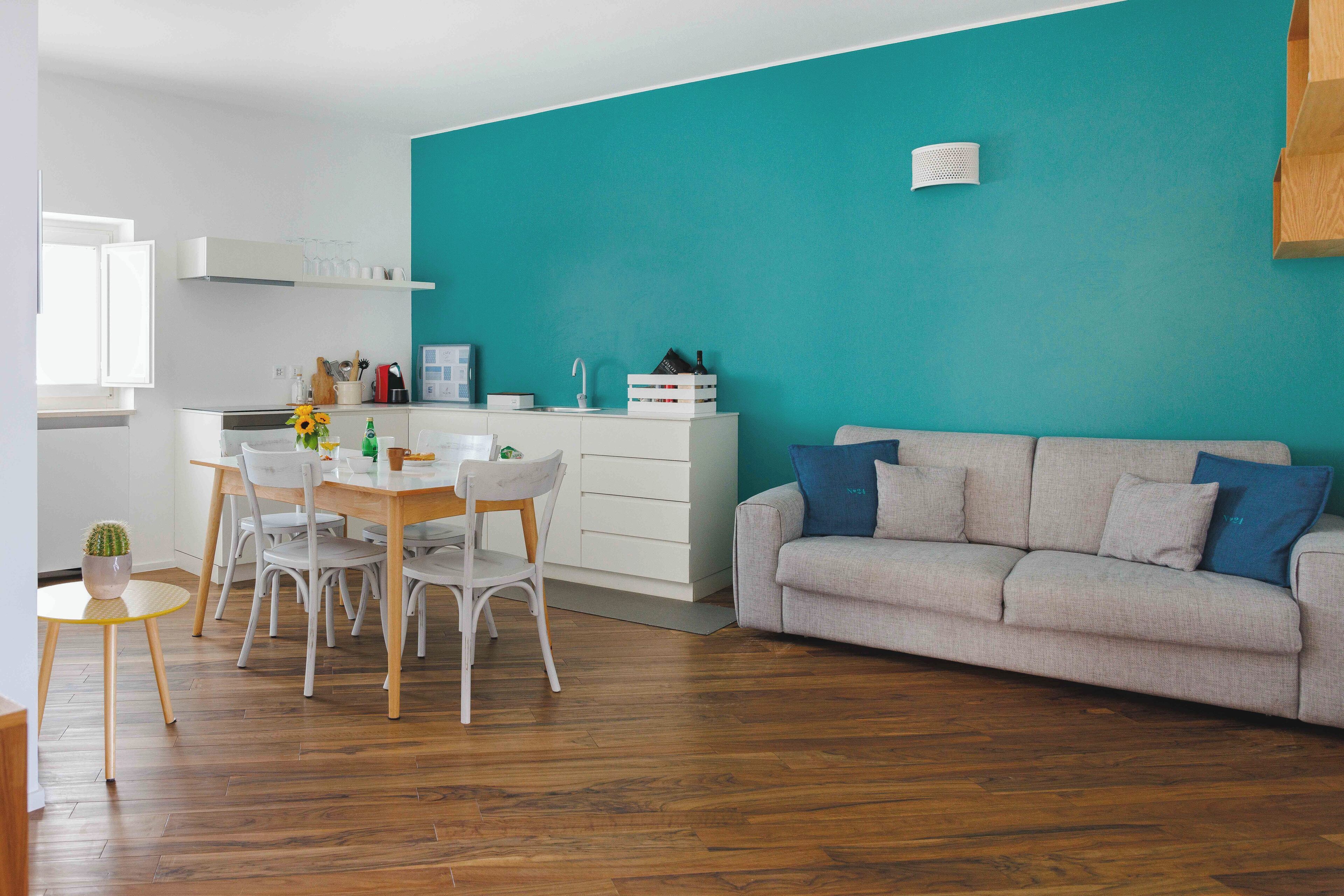 A modern living room with a teal accent wall, a grey sofa with blue cushions, and a wooden dining table with white chairs.