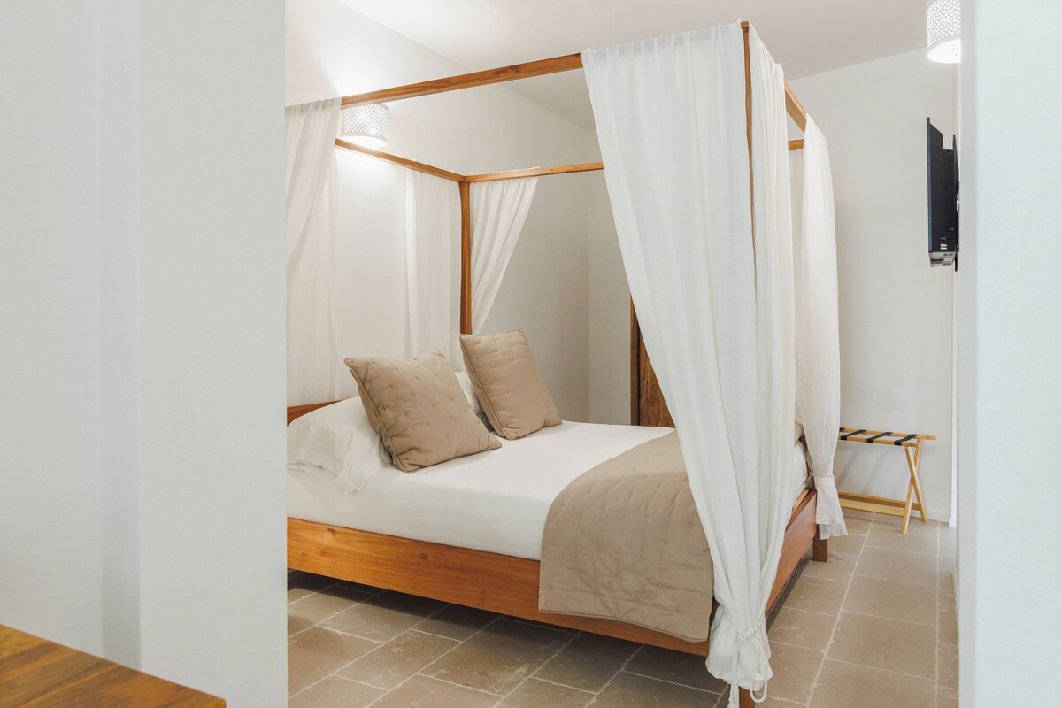 A four-poster bed with a wooden frame and white canopy, a small bedside table, and a wall-mounted TV.