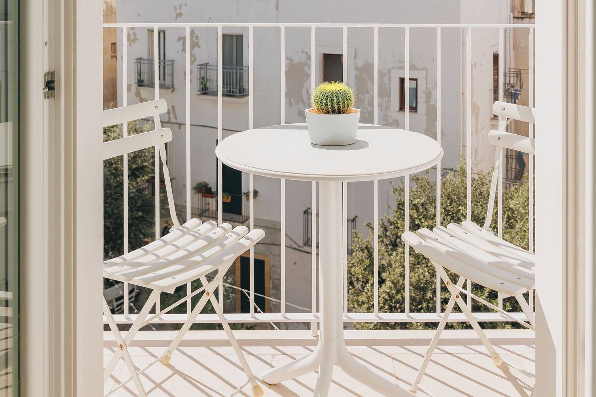 Un balcon avec une table et des chaises blanches, un cactus en pot et une vue sur des bâtiments historiques.