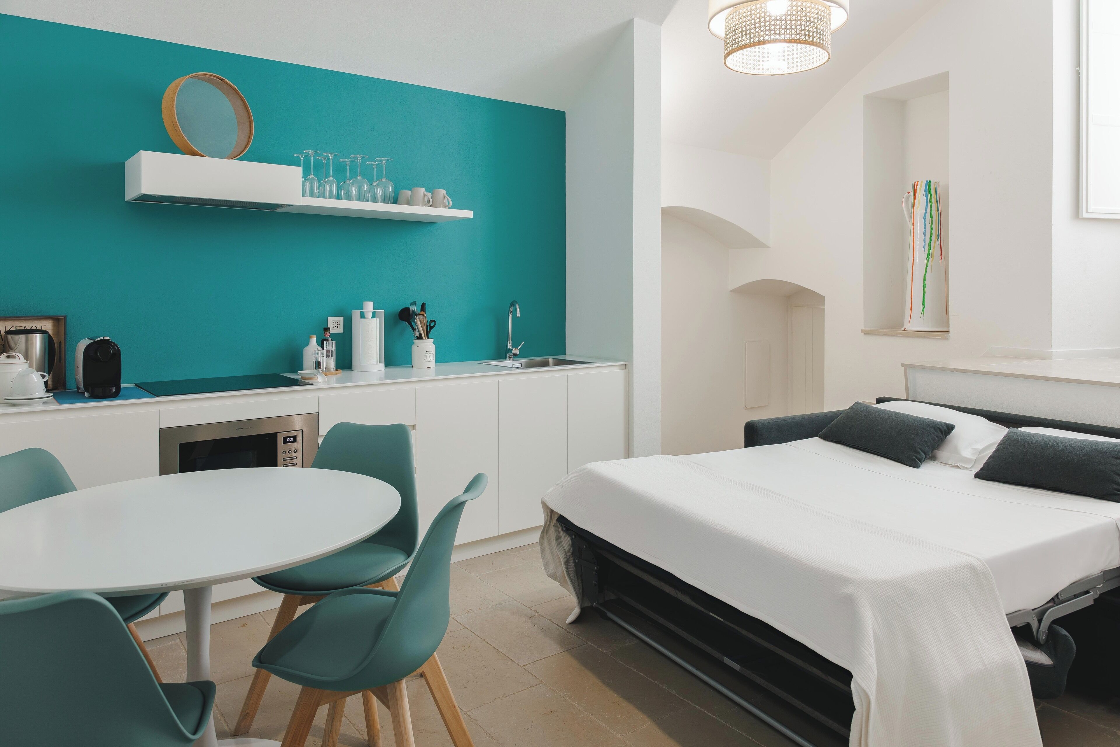 Una habitación moderna con una cama, una mesa de comedor con sillas, una zona de cocina con un lavamanos y una pared de acento color aguamarina.