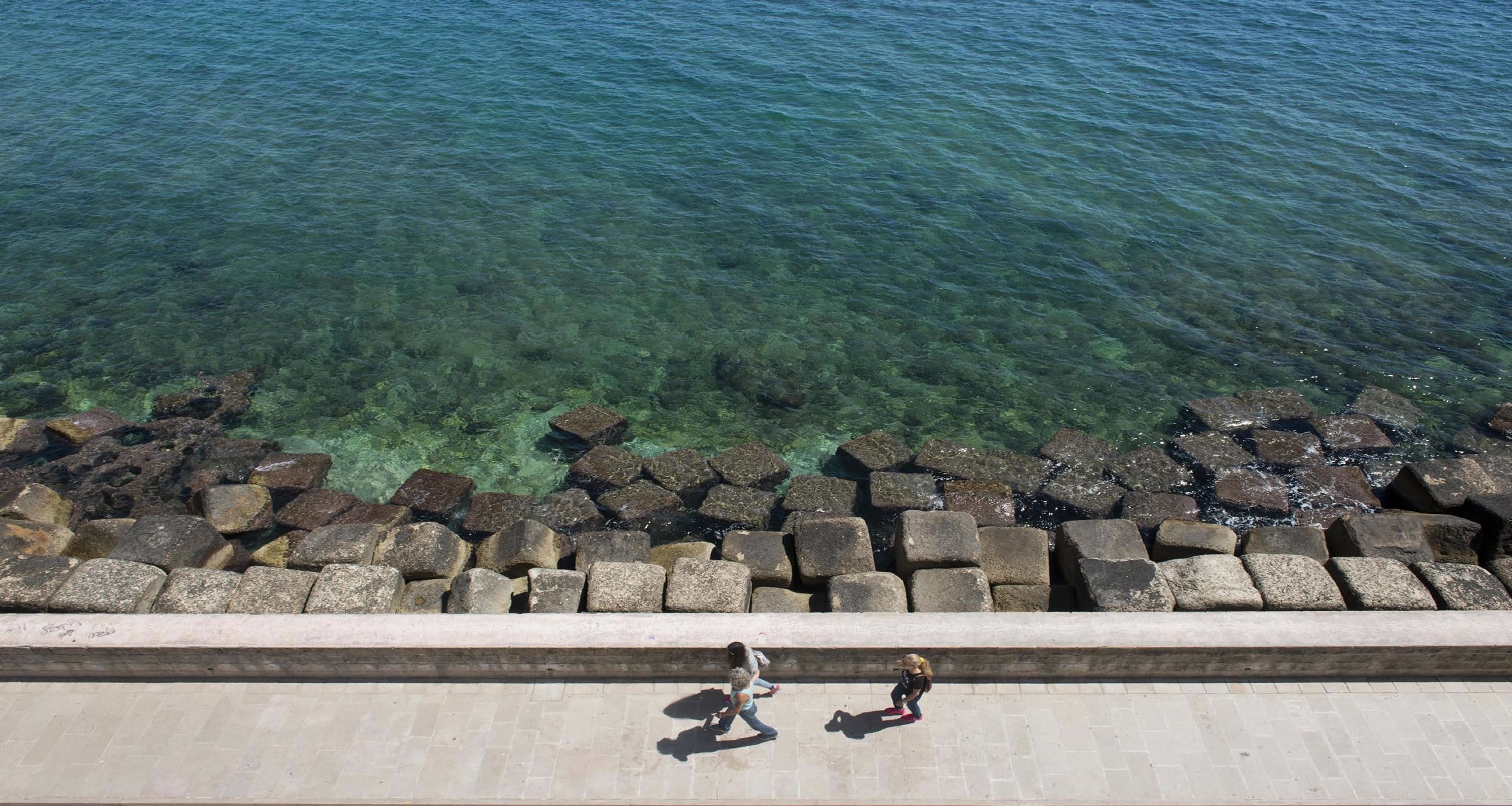 On voit clairement une promenade en bord de mer, avec une barrière en pierre et deux personnes qui marchent.