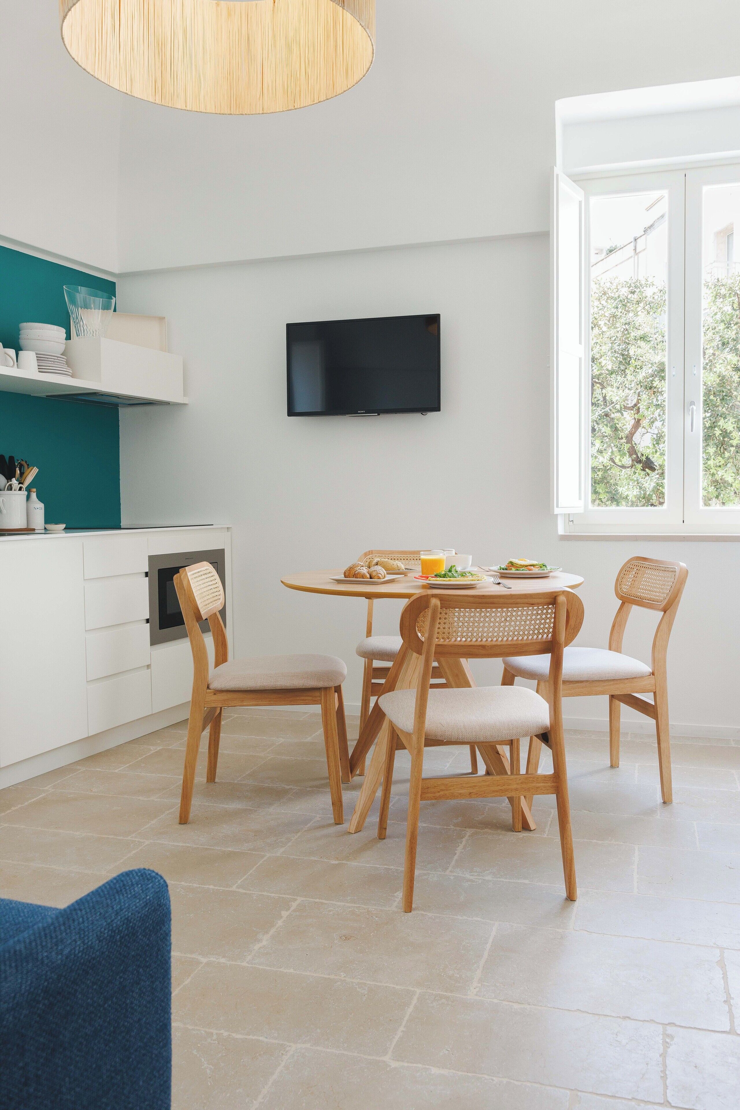 Une cuisine moderne avec des meubles blancs, un mur d’accent de couleur turquoise, un téléviseur à écran plat et un coin repas avec des chaises et une table en bois.