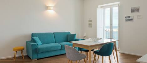 A modern living room with a teal sofa, a wooden dining table with chairs, and a small stool.