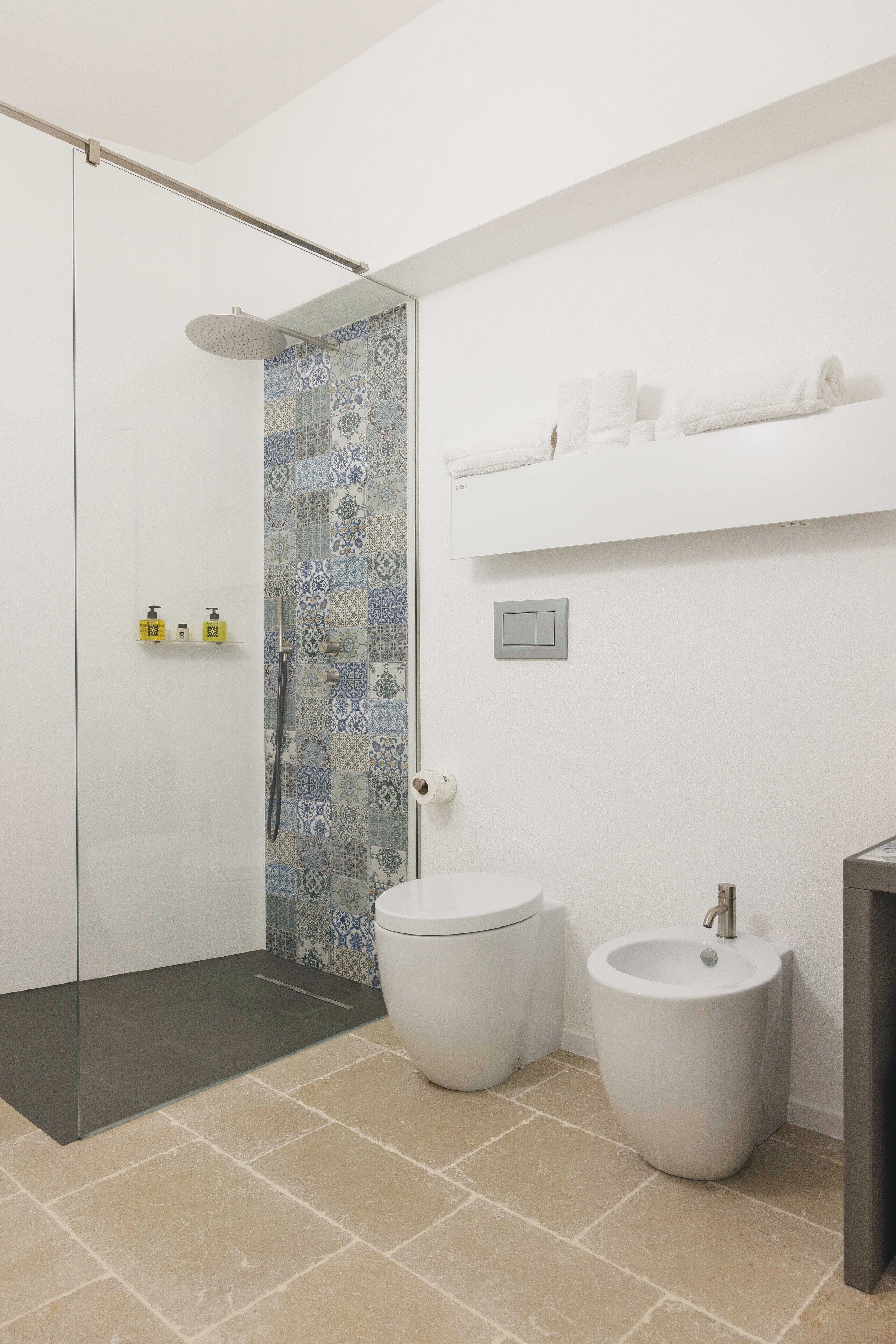 A modern bathroom with a glass shower enclosure, two toilets, a bidet, and a white shelf with towels.