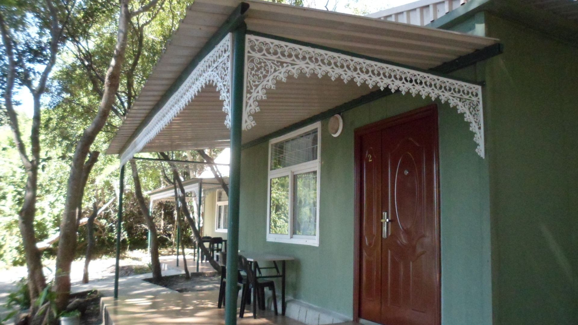 Un edificio con fachada verde, una puerta de madera y un porche cubierto con sillas.