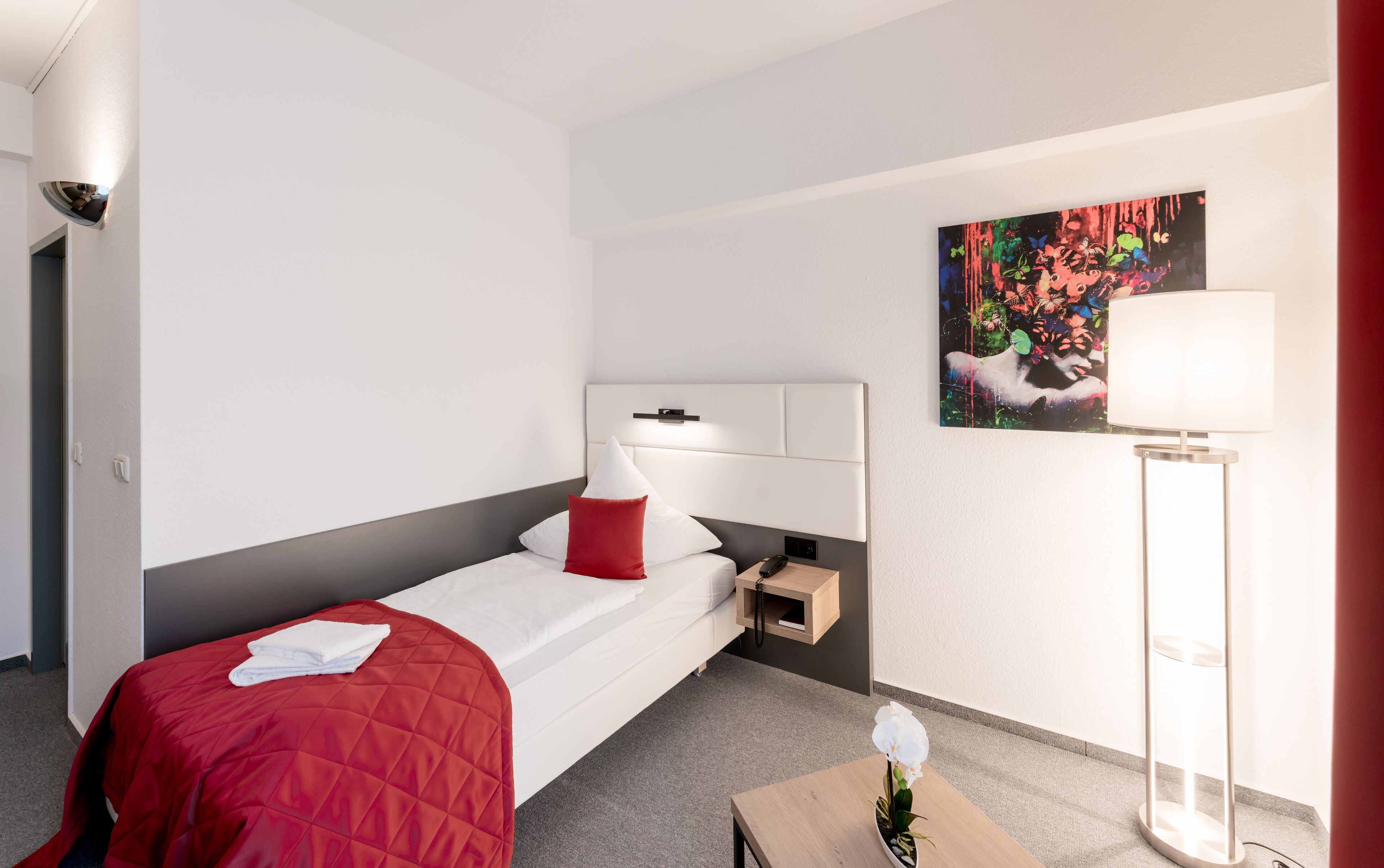 Habitación de hotel con una cama, una mesita de noche con lámpara, un cuadro en la pared y una planta pequeña en el suelo.