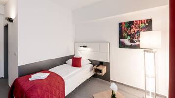 Habitación de hotel con una cama, una mesita de noche con lámpara, un cuadro en la pared y una planta pequeña en el suelo.