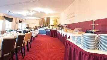 Une salle de banquet avec des tables rondes, un espace buffet et un sol recouvert d’un tapis rouge.