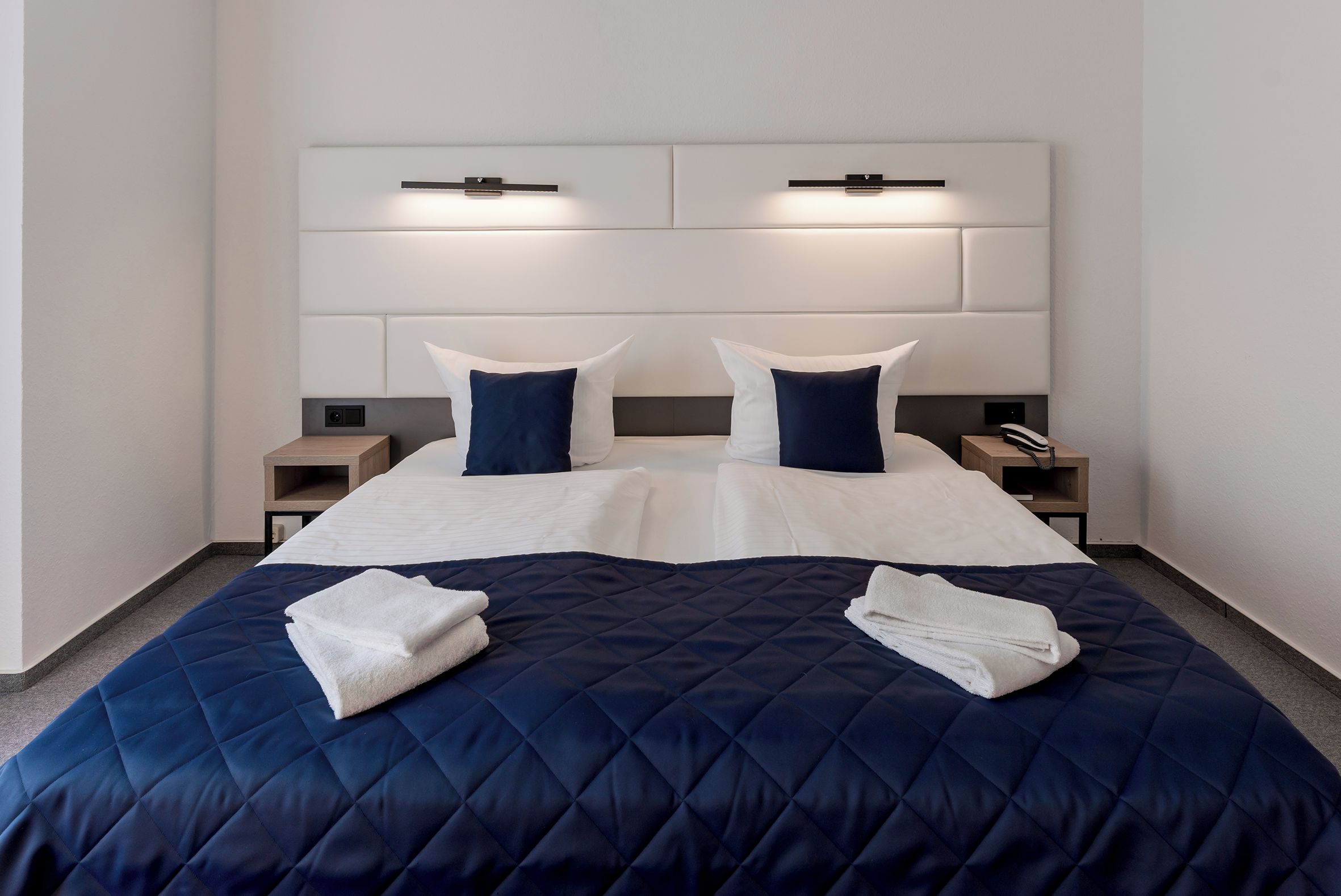 A hotel room with a large bed, two bedside tables, and a headboard with integrated lighting.