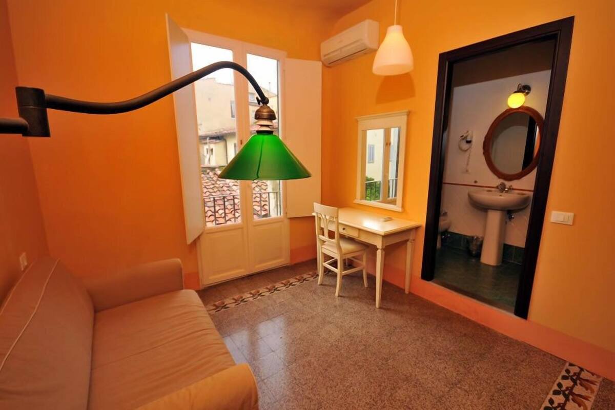 Et kompakt værelse med sofa, skrivebord, stol, vindue og badeværelse med spejl og håndvask.