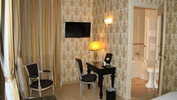 Habitación de hotel con cama, escritorio con lámpara, silla, televisión y baño visible a través de una puerta abierta.