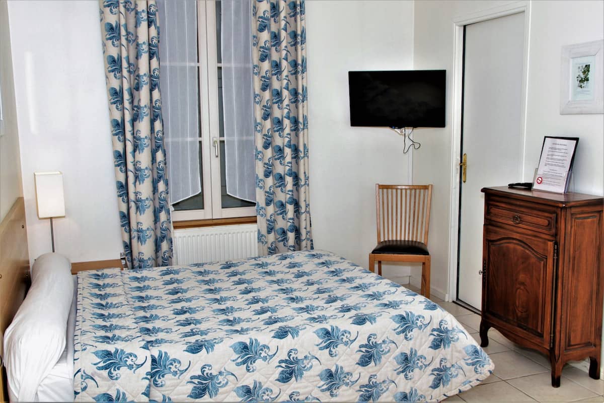 Ein Schlafzimmer mit einem Bett, einem Holzstuhl, einem Holzkasten, einem an der Wand befestigten Fernseher und einem Fenster mit Vorhängen.