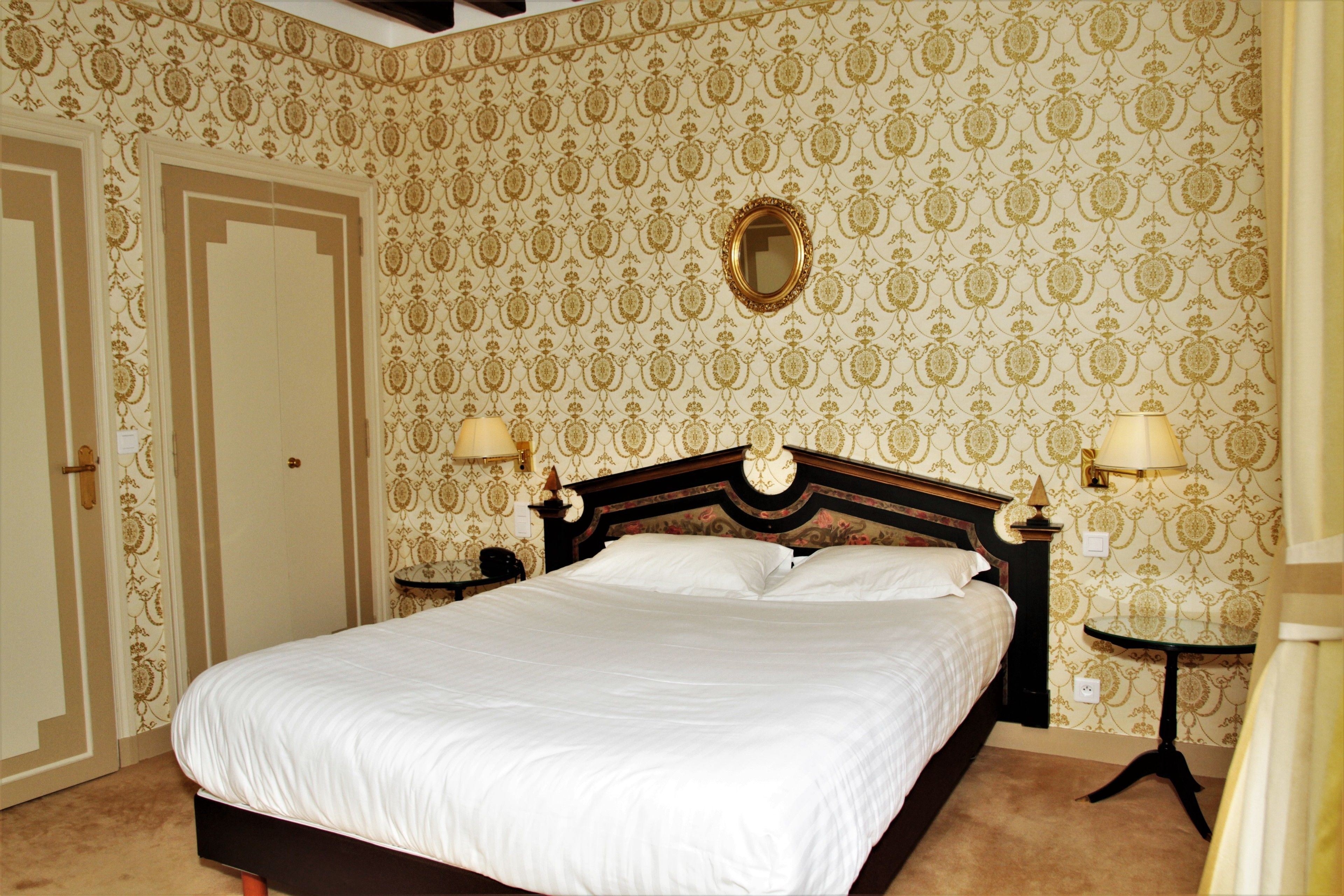 A neatly made bed in a room with patterned wallpaper, a mirror above the headboard, and two bedside lamps.