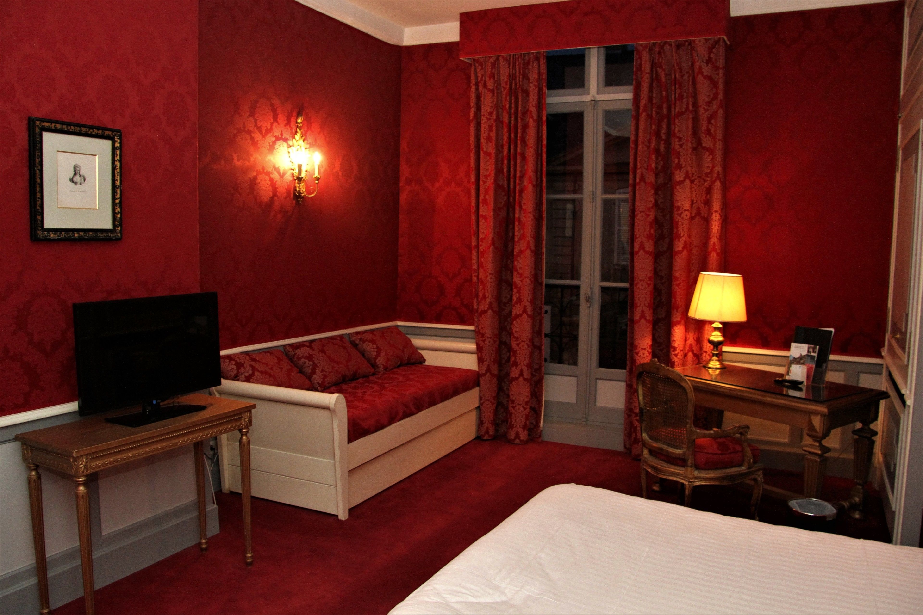 A hotel room with a bed, a television on a stand, a desk with a chair, and a window with curtains.