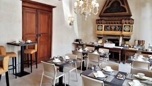 A dining area with multiple tables set for breakfast, a wooden door, and a decorative fireplace with heraldic shields. - Hotel Le Regent (Villers-Cotterets)