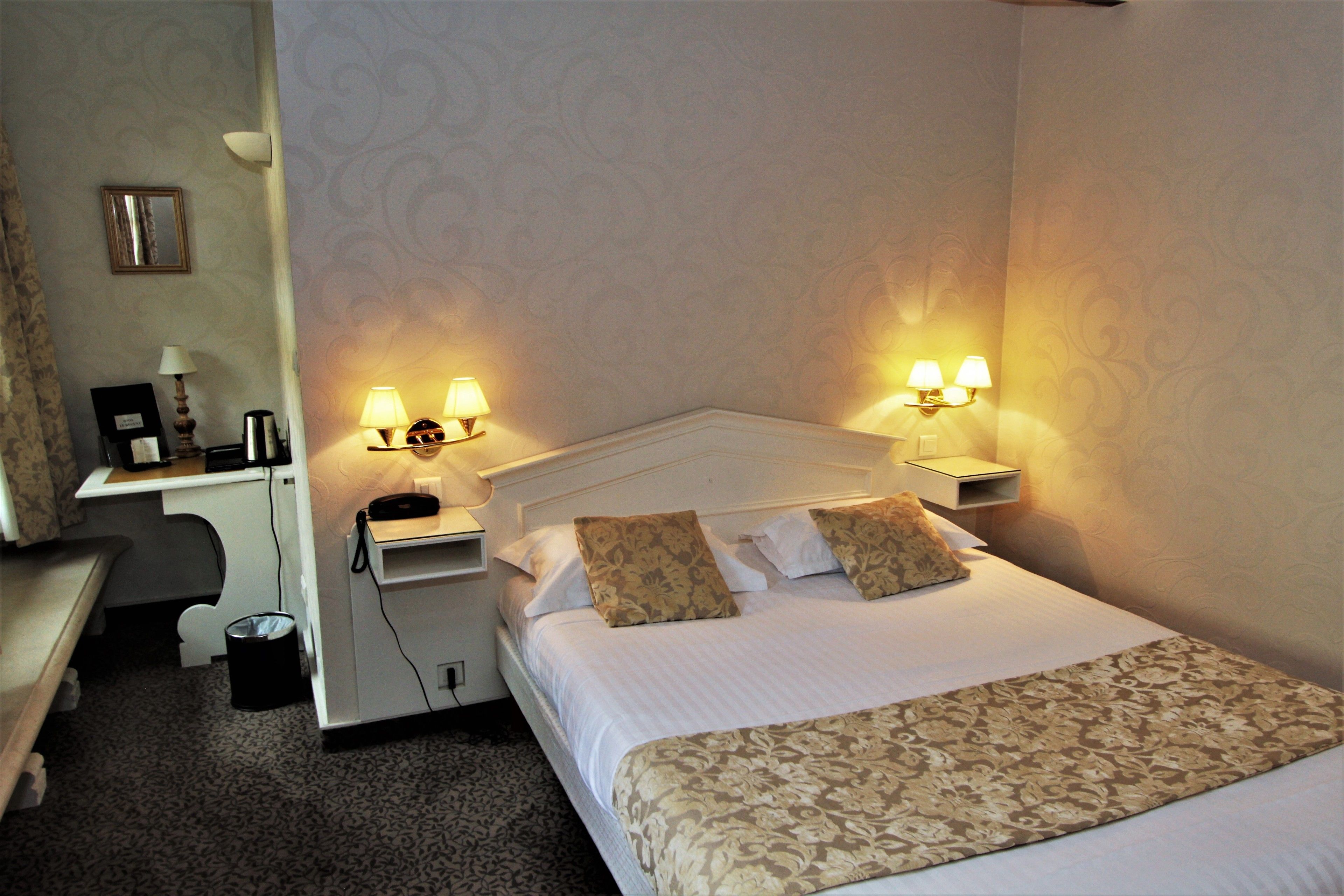 Une chambre d’hôtel avec un lit bien fait, deux lampes de chevet, un bureau avec une lampe et une petite poubelle.