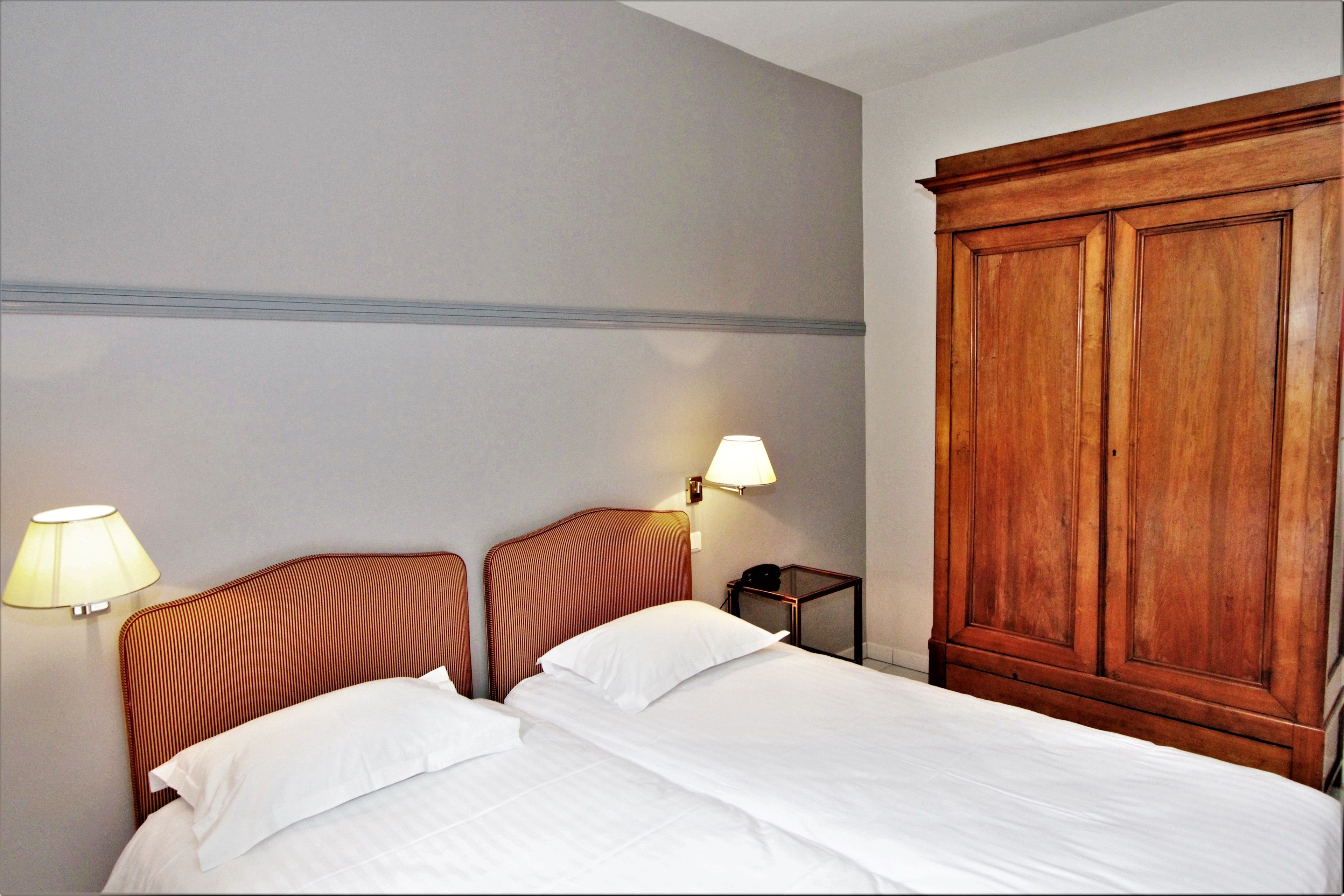 A hotel room with a large wooden wardrobe, a bed with white bedding, and two wall-mounted lamps.