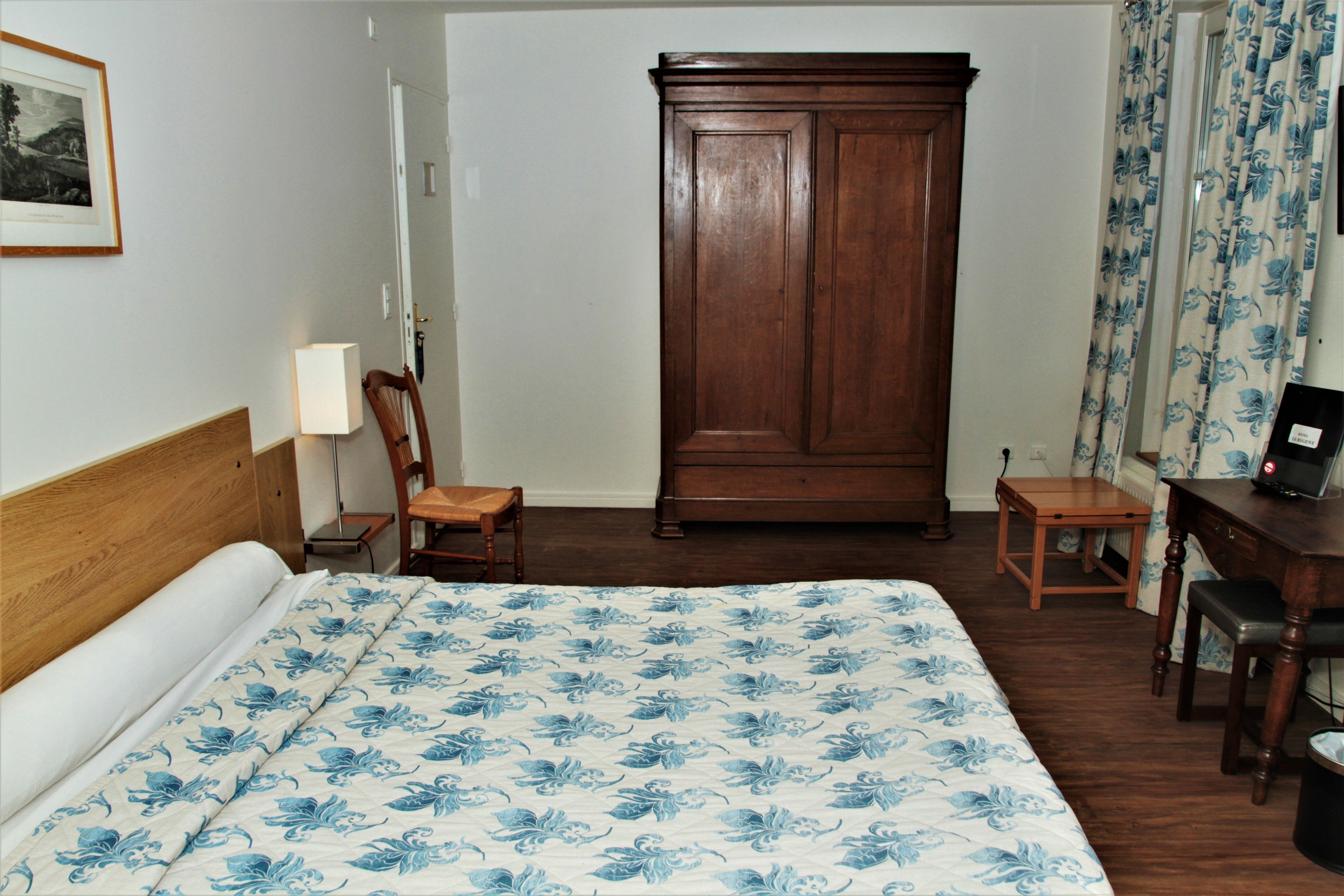 A hotel room with a large bed, a wooden wardrobe, a desk with a chair, a small table, and a lamp.