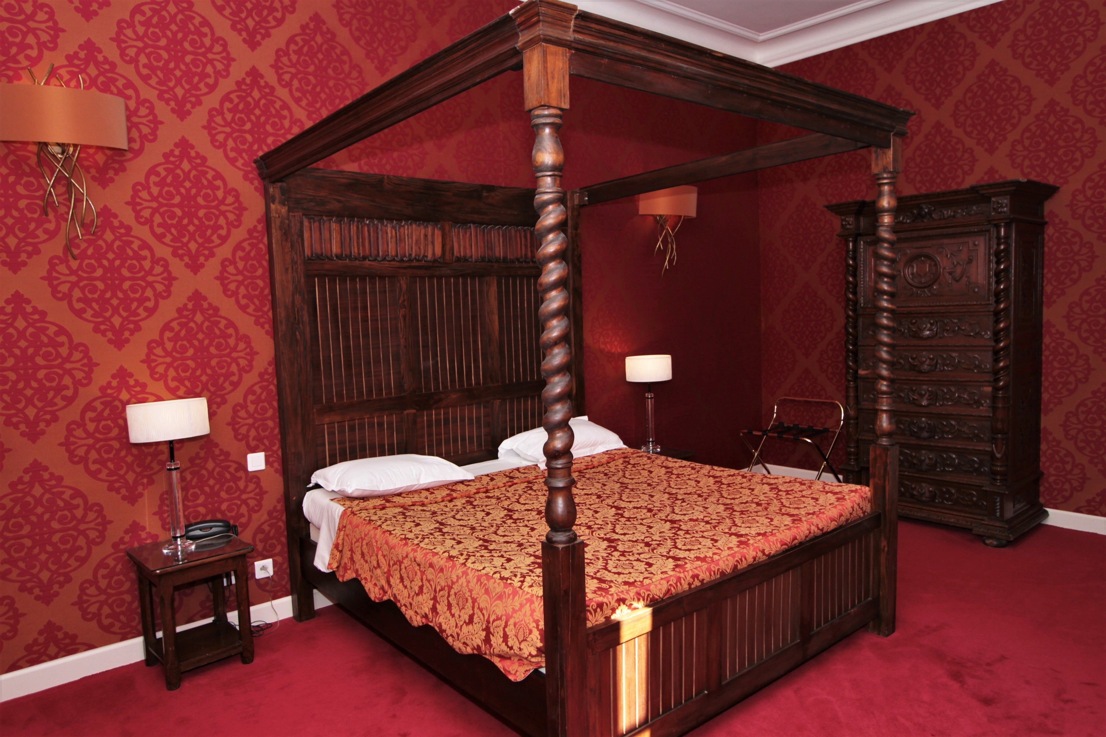Una cama con dosel de madera con un edredón rojo estampado, flanqueada por dos mesitas de noche con lámparas, en una habitación con papel tapiz rojo y un aparador de madera.