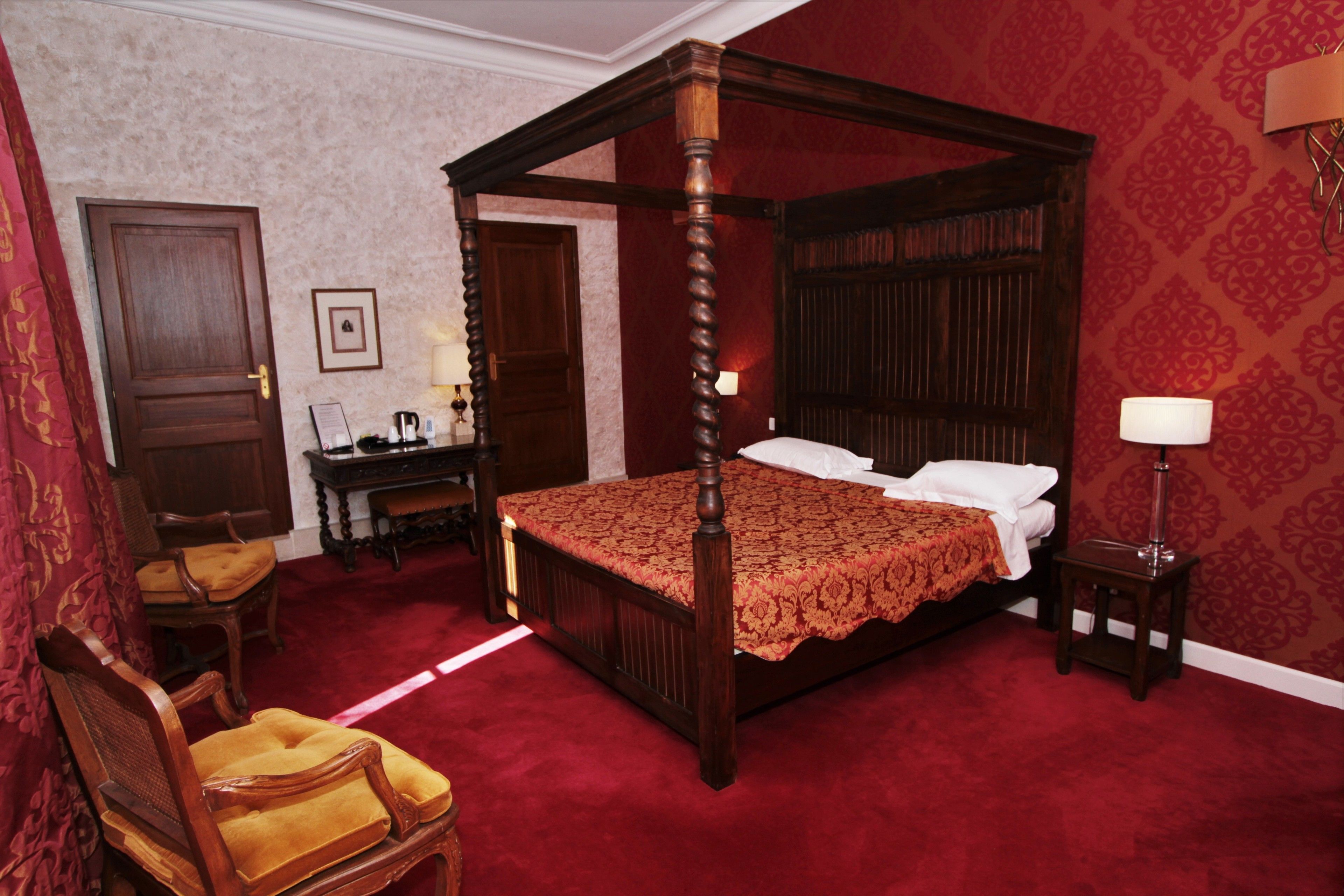 A four-poster bed with a red bedspread, a wooden nightstand with a lamp, a chair with a yellow cushion, and a desk with a lamp.