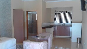 A modern living room with a sofa, a small round table, and a decorative chair. A kitchen area with a refrigerator and a window with striped curtains is visible in the background.