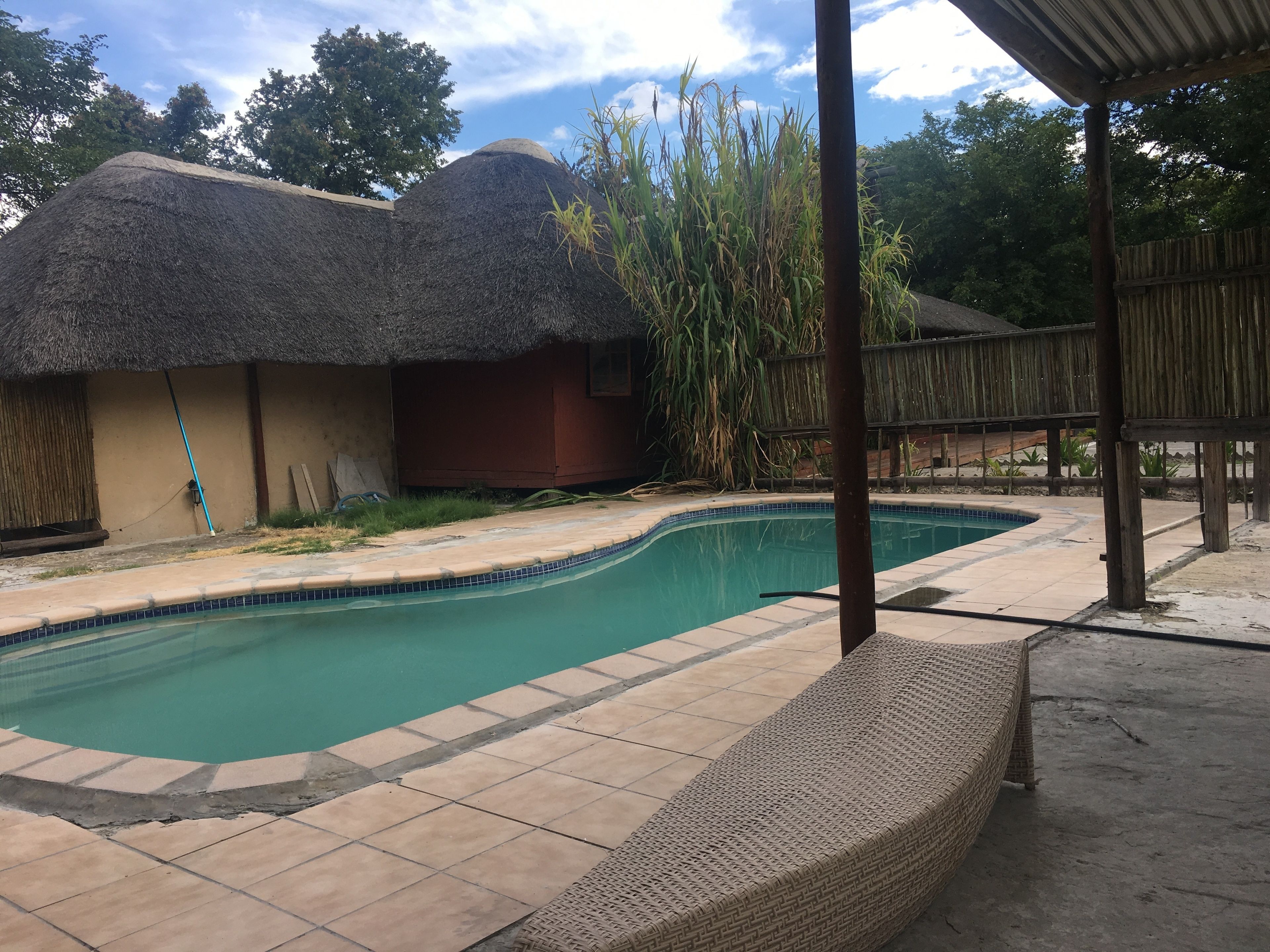 Ein Schwimmbecken mit gefliesten Rändern, ein Gebäude mit einem Reetdach und ein Holzzaun.